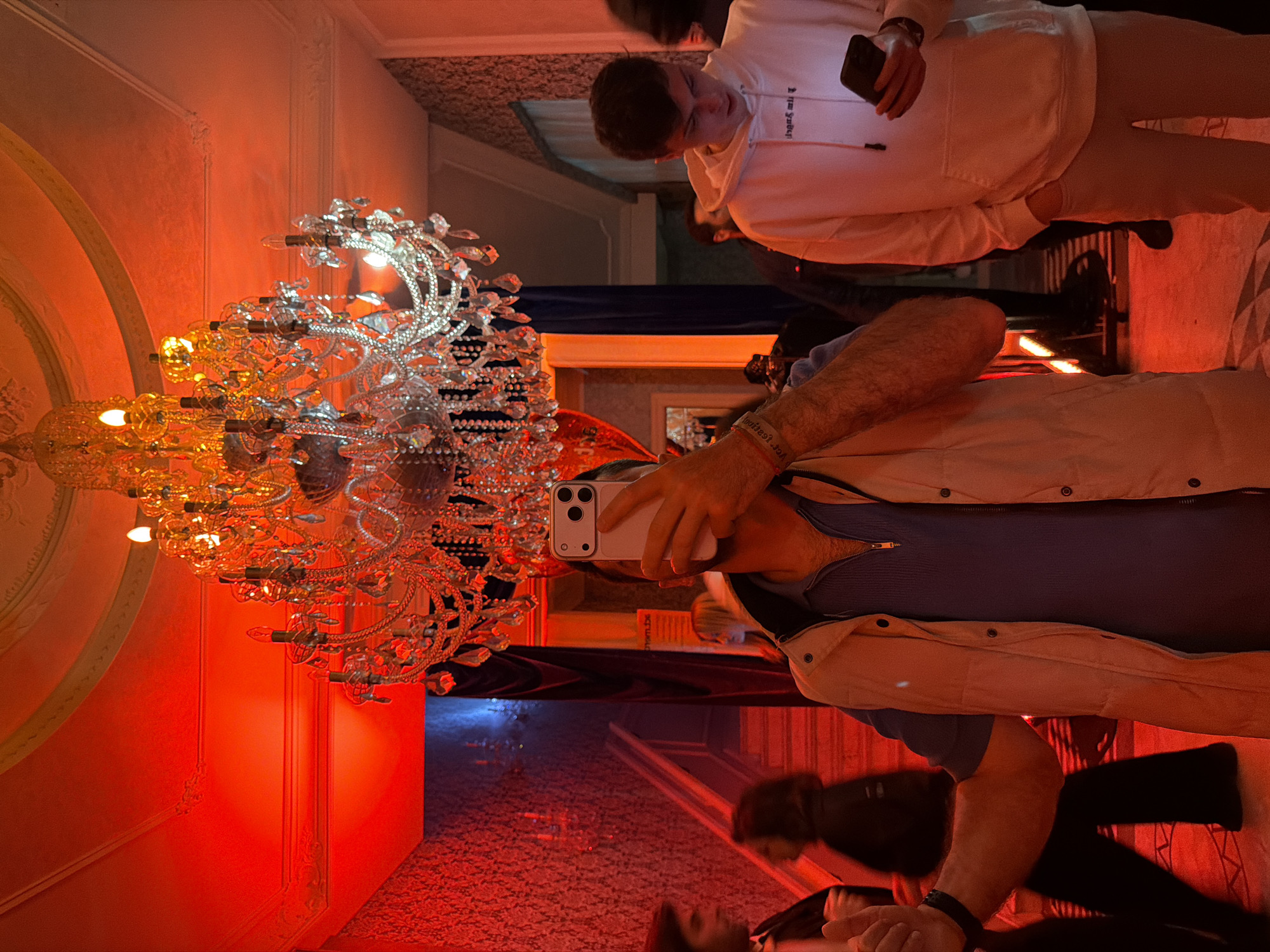The image shows an indoor scene in Tbilisi, Georgia, captured in portrait orientation. In the foreground, a chandelier with multiple light bulbs is positioned. A person wearing a beige vest over a blue shirt is seen taking a picture of the chandelier with a white smartphone. Another individual, dressed in a white hoodie and beige pants, also holds a smartphone. The setting appears to be an interior space with red walls, possibly a hotel or an upscale establishment. Reflections on a glossy surface add depth to the scene.