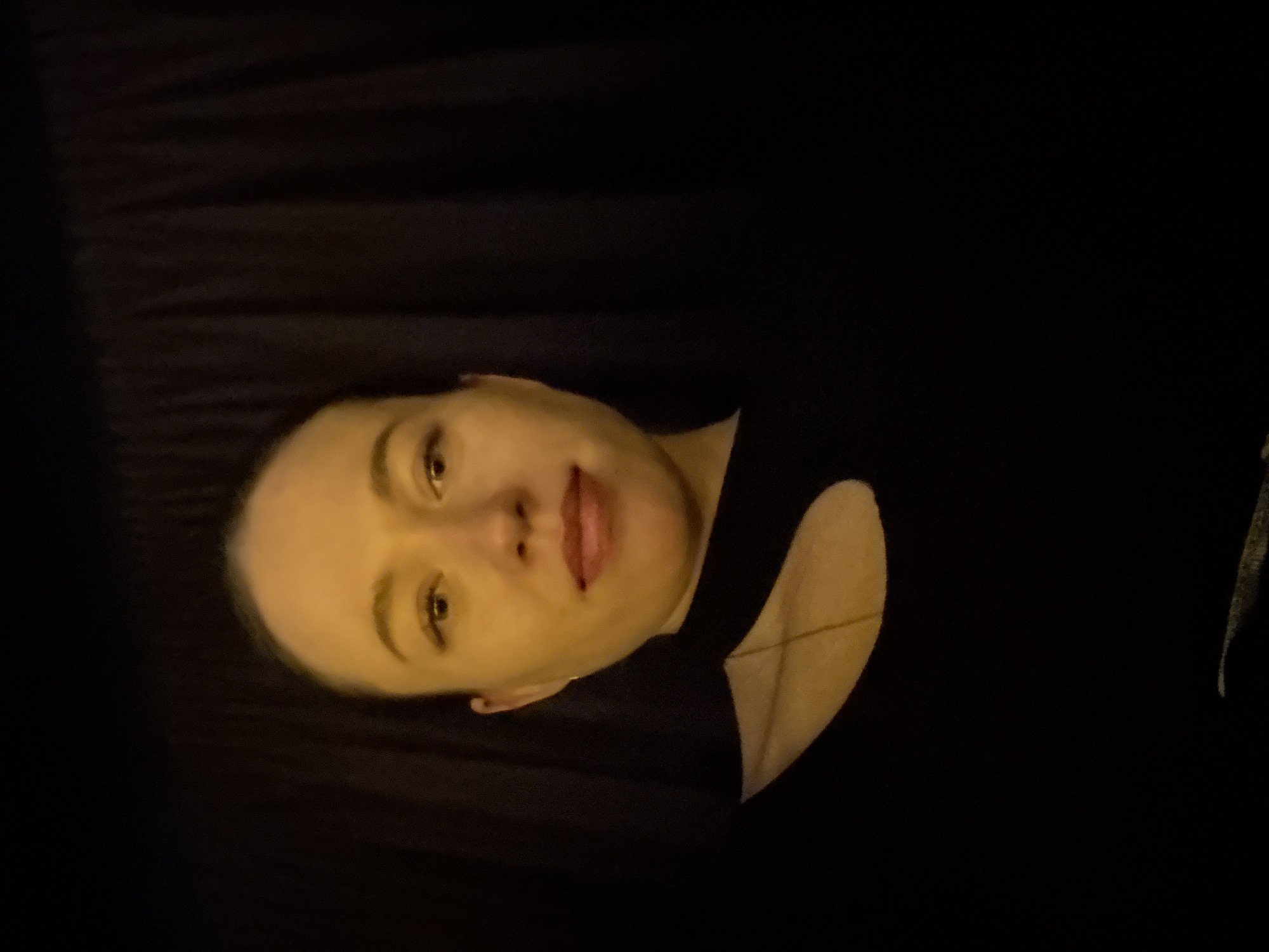 A woman with light skin is lying down on a dark surface that appears to be fabric. She is wearing a black top with a cutout on the shoulder. A delicate gold chain is visible around her neck. Her expression is neutral, and she is looking at the camera. The image has a slightly warm tone. The location in Perm, Russia is not discernible from the image.