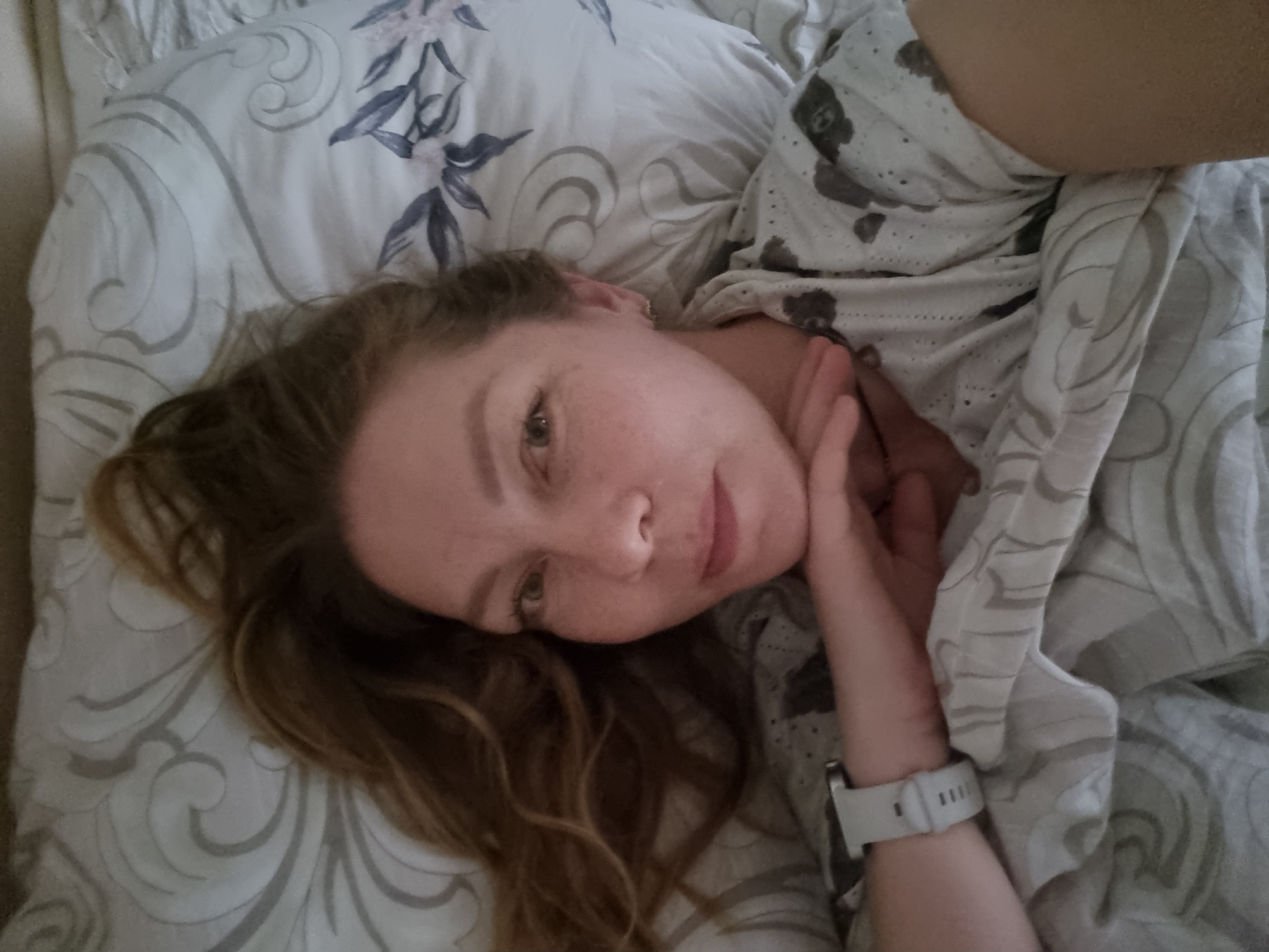 The image captures a woman, appearing to be in her 20s or 30s, lying in bed and looking directly at the camera. Her wavy, light brown hair is spread across the pillow. She has visible freckles on her nose and cheeks, and her expression is neutral and serene.
She is partially covered by light-colored bedding, which appears to be a duvet or blanket. This particular piece of bedding has a subtle pattern of small, dark motifs resembling teddy bears and a textured finish, possibly eyelet. Her head rests on a pillow that features a gray and white design with both floral or leaf elements and swirling patterns. Another visible section of bedding also displays a similar gray swirling pattern. A light gray or white smartwatch with a silicone strap is worn on her left wrist.
The scene is set indoors, likely a bedroom, suggesting a moment of relaxation or rest. The lighting is soft and diffused, indicating it could be morning, late afternoon, or early evening. The woman's direct gaze suggests she might be taking a selfie or posing for a close-up photograph. There is no visible text in the image. This indoor scene is situated in Chaikovsky, Russia.