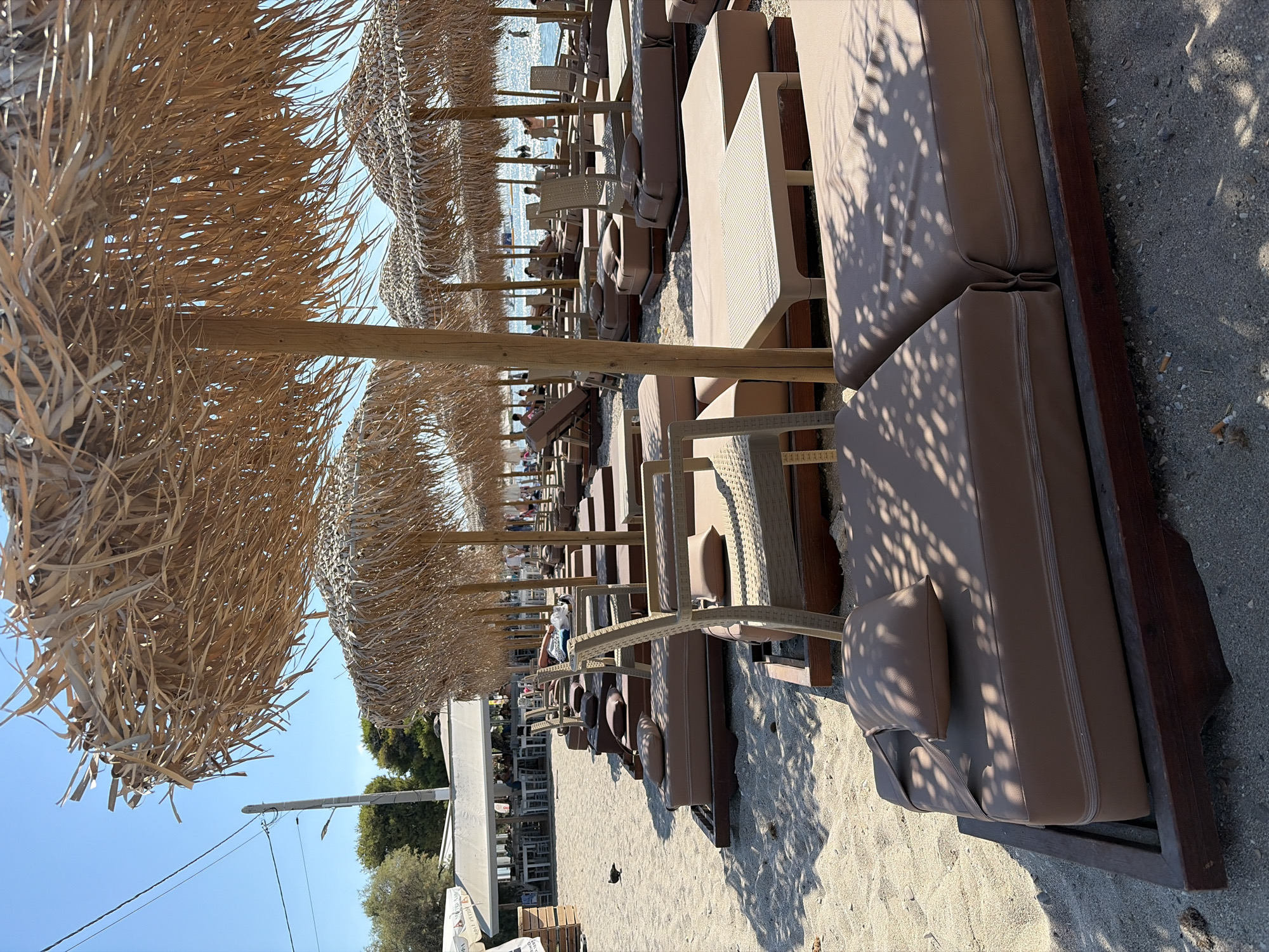 This image depicts a beach with rows of empty, cushioned loungers and thatched umbrellas. The loungers are arranged in neat rows on a sandy beach, and several small tables are placed between them. In the background, a beach bar or restaurant with outdoor seating is visible, and a few people are seen walking near the water. The sky is clear and blue. The scene suggests a peaceful, relaxed atmosphere on a sunny day.