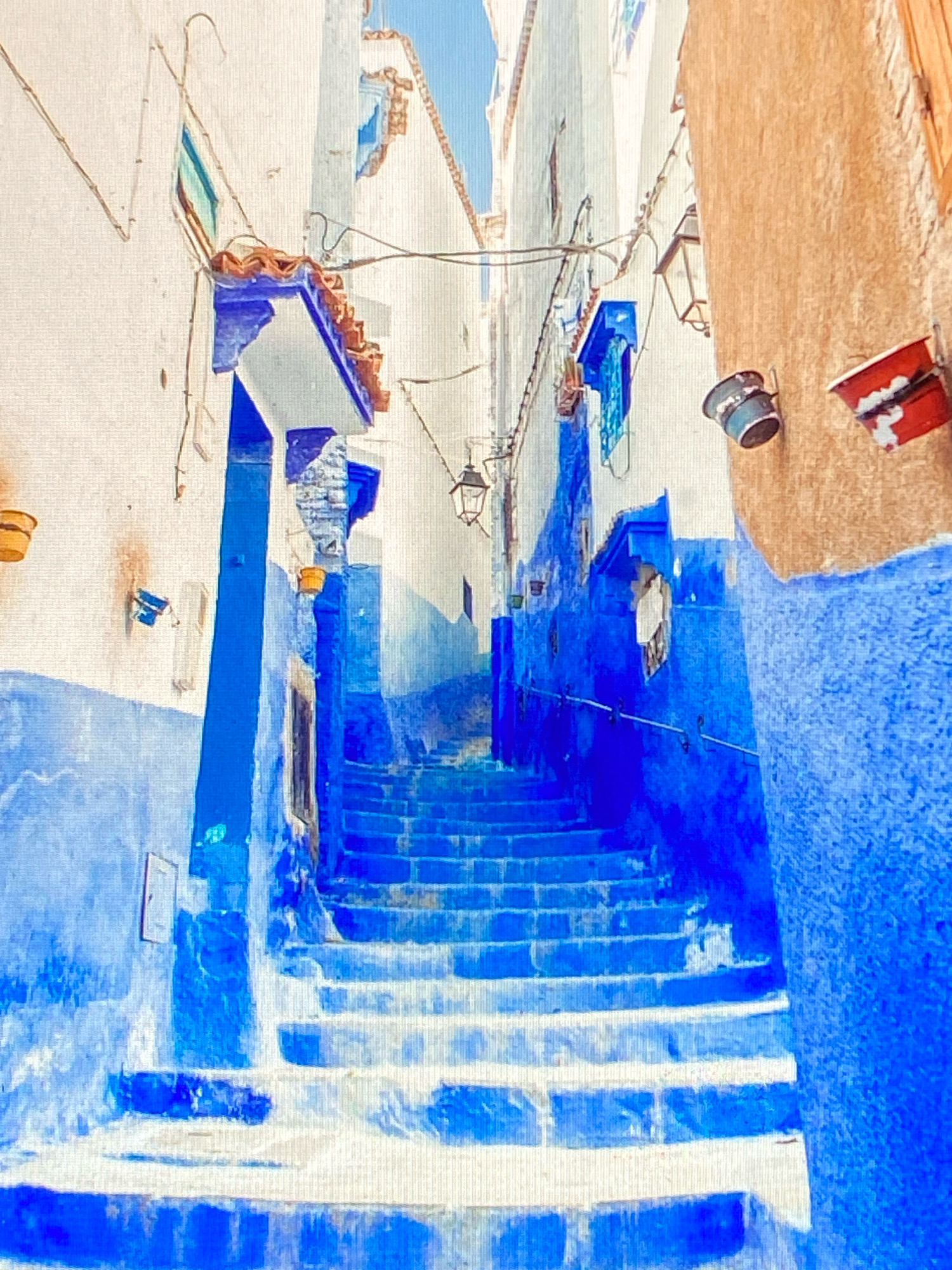 The image shows a narrow alleyway in the city of Salé, Morocco, characterized by its vibrant blue-painted walls and stairs. The alley ascends into the distance, with buildings on either side. The architecture features white-washed walls with blue accents around windows and doorways. Terracotta-tiled roofs are visible. Several decorative elements are present, including hanging lanterns, potted plants, and what appear to be repurposed containers painted red and blue mounted on a textured, ochre-colored wall on the right. The ground is covered by a series of blue and white steps. The scene is devoid of any people. The lighting suggests it is daytime, with a clear sky visible at the top of the alley.