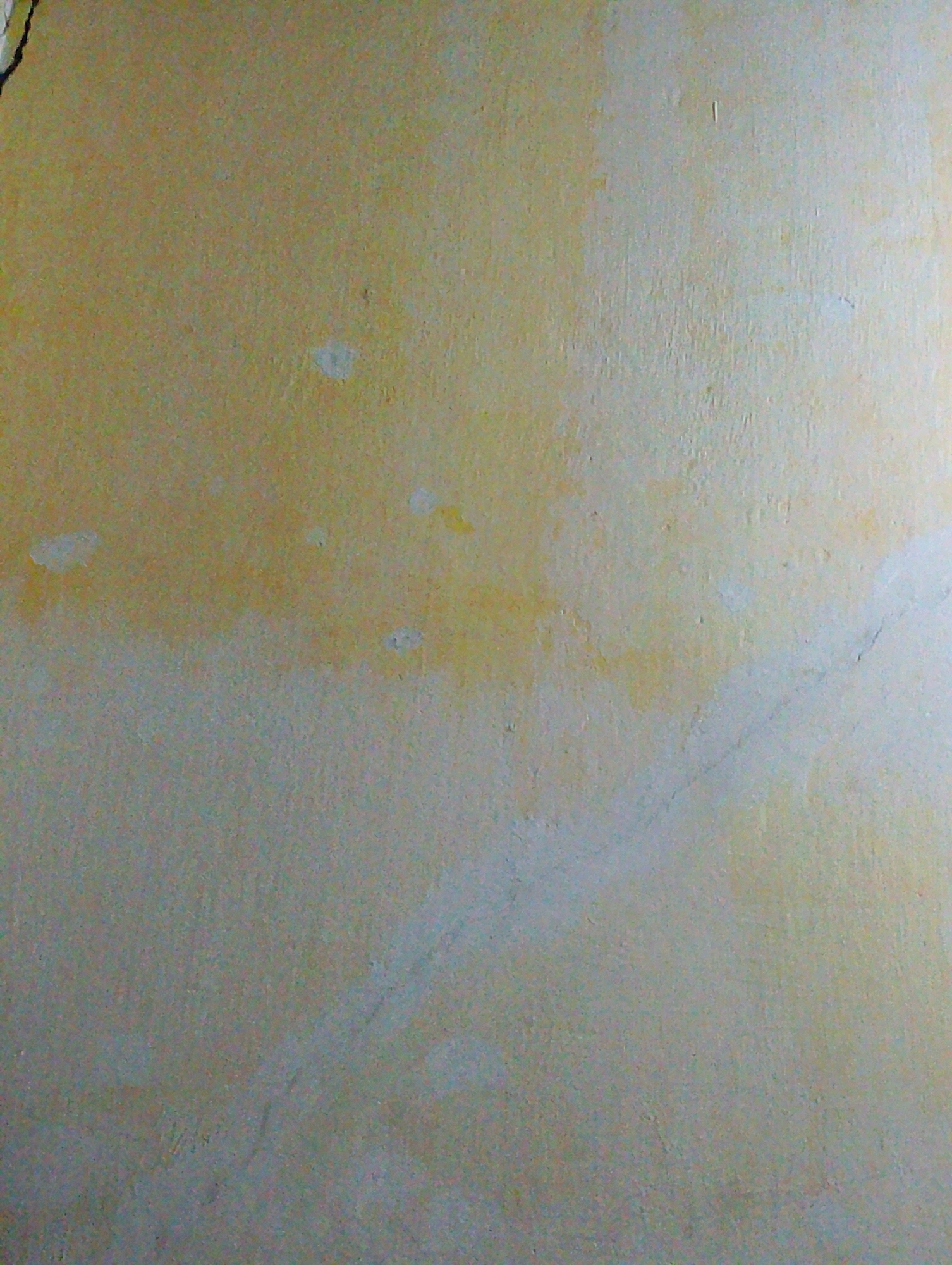 The image displays a textured, discolored wall. The upper portion of the wall exhibits a yellowish hue with irregular patches of lighter color and subtle staining. As the wall extends downwards, the color transitions to a lighter, more muted tone, with faint gray and blue undertones. The surface appears rough and uneven, with visible imperfections and possibly some fine cracks. No people, objects, or discernible actions are present. The location within the city of Plosoklaten, Indonesia, is not identifiable from the visual information provided in the image.