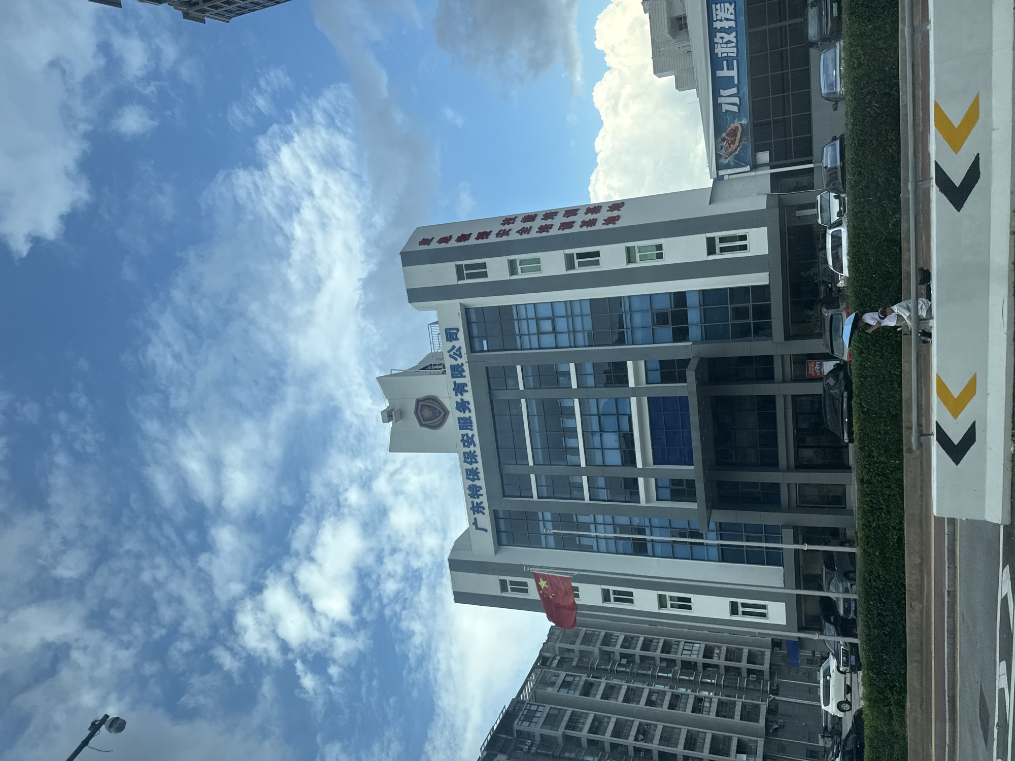 The image shows a modern, multi-story building in Huizhou, China. The building has a grey and white facade with blue-tinted windows. A Chinese flag is prominently displayed on a flagpole to the left of the main entrance. Text in Chinese characters is visible on the upper portion of the building, appearing to be signage for a security service company and a training base.
In the foreground, a road with traffic lane markings is visible. Several cars are parked in front of the building. A person is standing on a sidewalk with an umbrella, and another person is seated on the ground nearby. The sky is blue with scattered white clouds. To the left of the main building, a taller, grey, residential building can be seen. To the right, a blue banner with Chinese text and an image of a life raft is attached to the building.