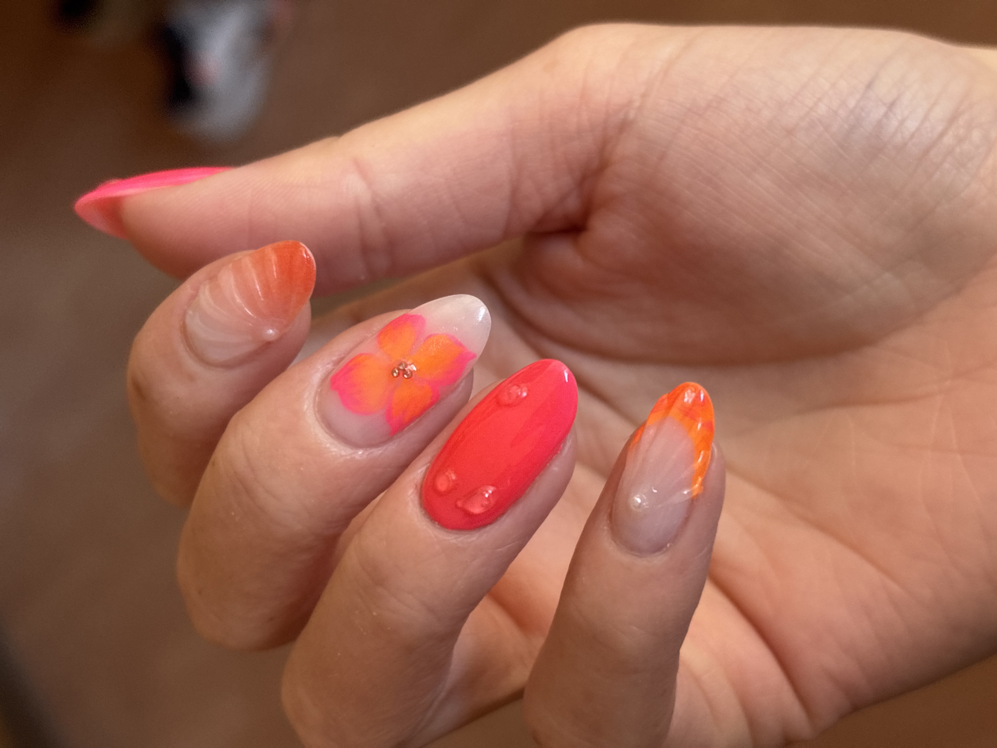 A close-up shot features a person's left hand, adorned with an elaborate and colorful manicure. The hand is positioned with fingers slightly splayed, showcasing each nail.
Each nail displays a distinct design, predominantly in shades of pink, orange, and red-orange with clear bases. The nail on the thumb, partially visible on the far left, is painted a solid, bright pink-orange. The index finger features a clear base with an ombre or gradient effect, transitioning from a clear base to a vibrant orange-pink tip. The middle finger showcases a delicate floral design on a clear base, with petals in orange and pink hues, centered by three tiny, golden spherical embellishments. The ring finger is painted a solid, bright coral red-orange and is adorned with multiple clear, raised droplets, simulating water drops. The pinky finger has a clear base with a vibrant orange tip in a classic French manicure style.
The nails are uniformly shaped in an almond or oval style. The background is softly blurred, showing warm, muted brown and beige tones, suggesting an indoor setting. The lighting is warm and even, highlighting the intricate details and glossy finish of the nail art. There is no visible text in the image.