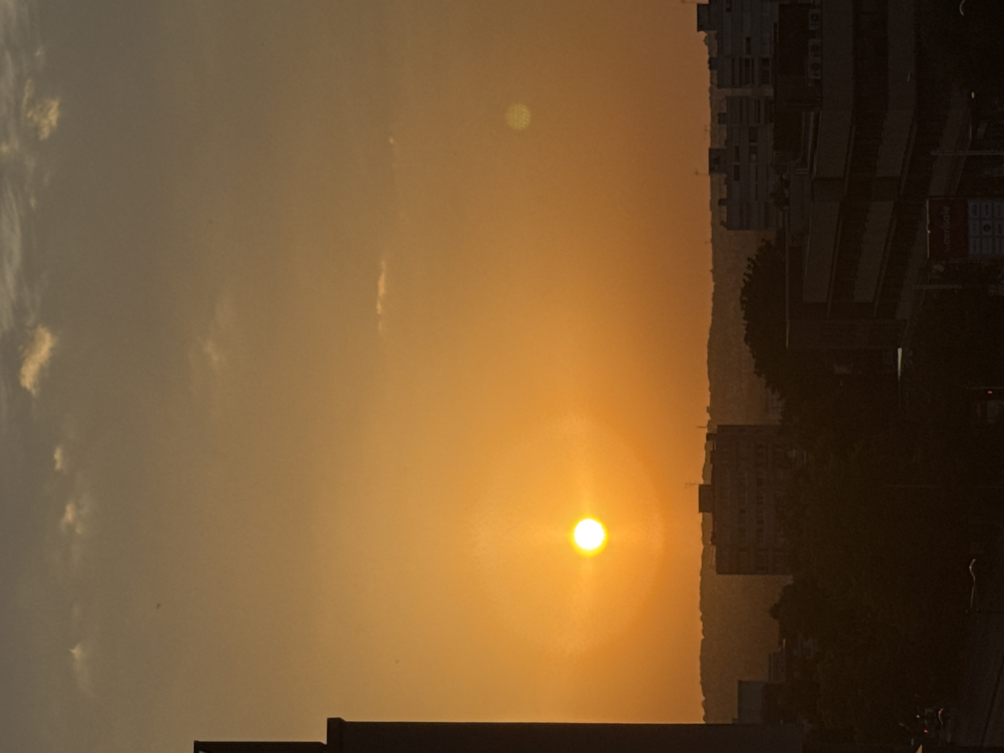 The image depicts a city skyline at sunset. The sun, a bright orange orb, is low in the sky and casts a warm, golden glow across the scene. A tall, dark building is silhouetted on the left side of the frame, obscuring part of the sunset. In the mid-ground, a cluster of buildings, likely part of the urban landscape, are visible against the hazy horizon. Some of these structures appear to be residential or office buildings, with visible windows and architectural details. Dark silhouettes of trees are present in the foreground, adding depth to the composition. The sky is a gradient of orange and yellow, with a few wispy clouds scattered across it. The overall impression is one of a tranquil evening in an urban environment.