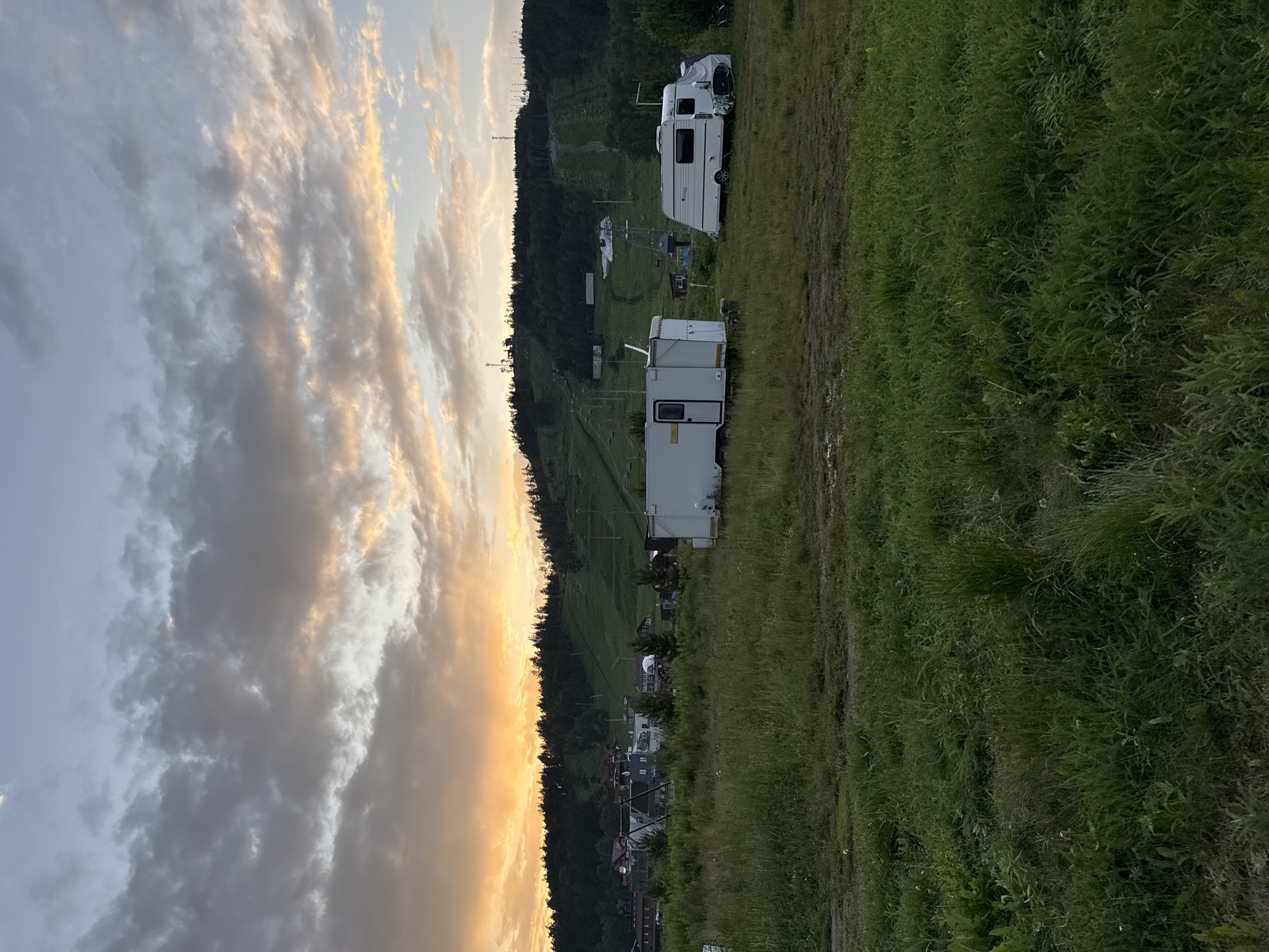 This image depicts an outdoor scene at dusk in Syrostan, Russia. In the foreground, a grassy field with tall green grass extends towards the middle ground. Two white recreational vehicles, likely campers or trailers, are parked in the field. The one on the left is smaller and boxier, while the one on the right is larger and more elongated, resembling a caravan.
In the middle ground and background, there is a cluster of buildings, possibly houses or cabins, nestled amongst trees. Behind the buildings, a sloping green hill rises, featuring ski slopes with visible ski lifts and poles. The hill is covered in lush green vegetation and appears to be part of a recreational area. The horizon is lined with a dense forest of dark green trees.
The sky above is a dramatic display of sunset colors. The upper portion of the sky is a pale blue, transitioning to shades of gray and pink as it nears the horizon. Streaks of vibrant orange and yellow light emanate from the setting sun, illuminating the scattered clouds and creating a warm glow. The overall impression is a tranquil, rural landscape at the end of the day.
