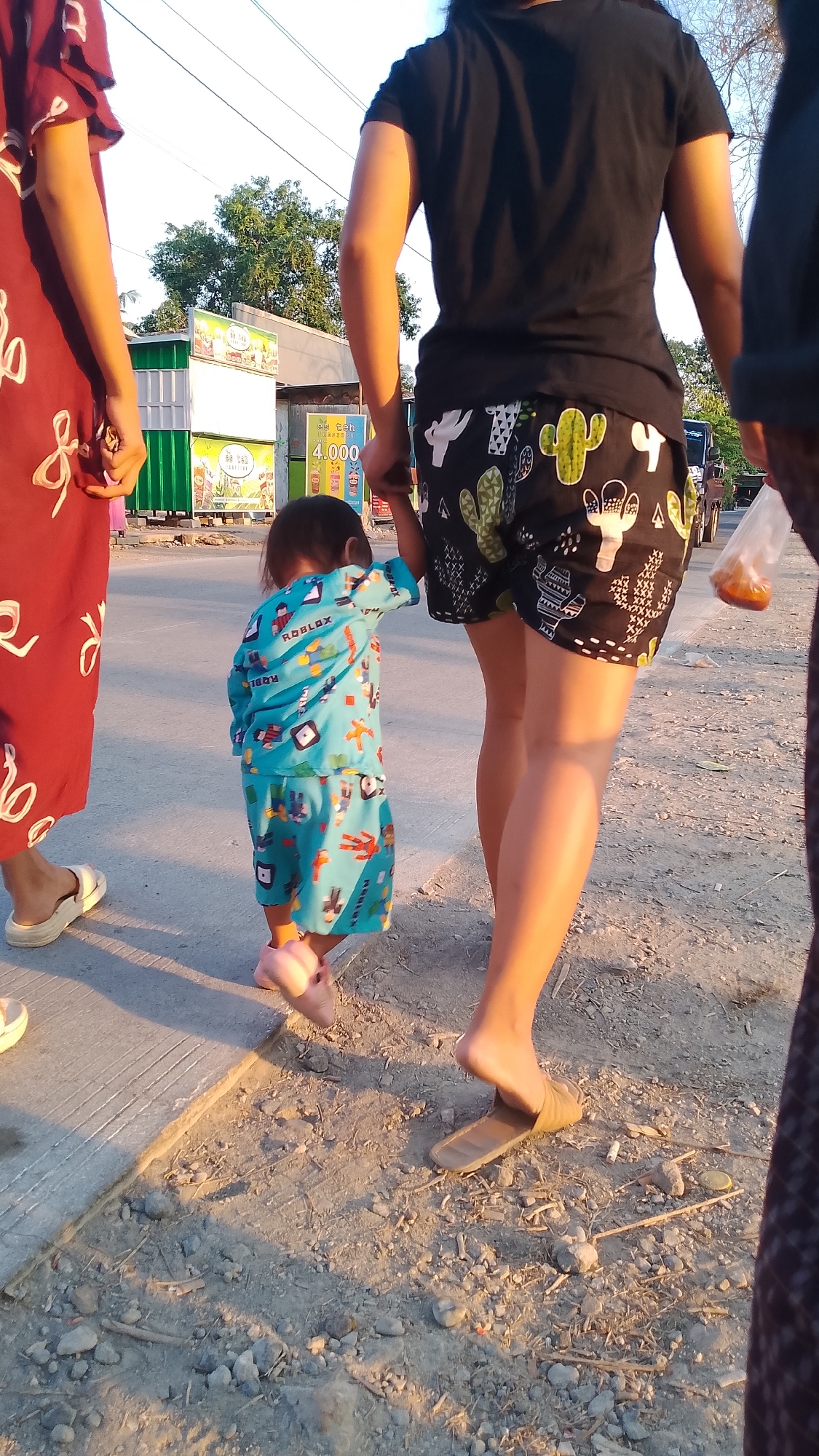 The image captures a rear view of three individuals walking along a dirt path next to a paved area. The person on the left, partially visible, wears a long, dark red dress with white patterns and white sandals. In the center, a young child, seen from behind, is dressed in a light blue pajama set with a Roblox theme and pink shoes. The child is holding hands with the person on the right. This individual, a woman, is wearing a black t-shirt and black shorts with a cactus pattern. She is also wearing brown slide sandals and is holding a clear plastic bag containing orange food. In the background, there are several small structures, one of which is a food stall with signage indicating "Es Teh 4.000". Trees and foliage are visible behind the structures. The scene appears to be an outdoor setting, possibly a street or market area in Karanglangu, Indonesia, as indicated by the presence of the food stall and the general environment. The lighting suggests it is late afternoon or early evening.
