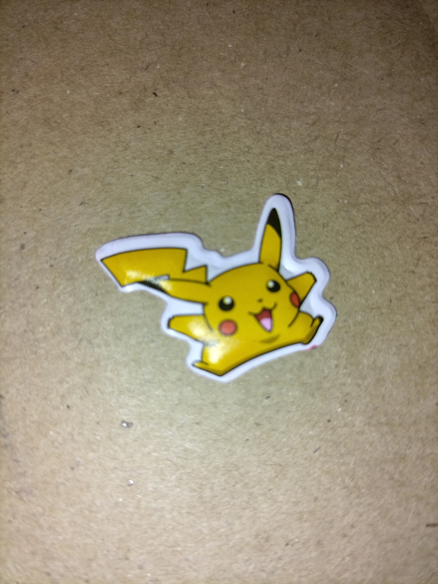 A Pikachu sticker is the sole subject of the image. The sticker is yellow with black and red accents and a white outline. It is positioned on a textured, brown surface that appears to be cardboard or a similar material. The sticker is at a slight angle and appears to be a sticker cut out in the shape of Pikachu. There are no people, city elements, or specific locations visible in the image. The lighting is dim and slightly casts shadows on the sticker.