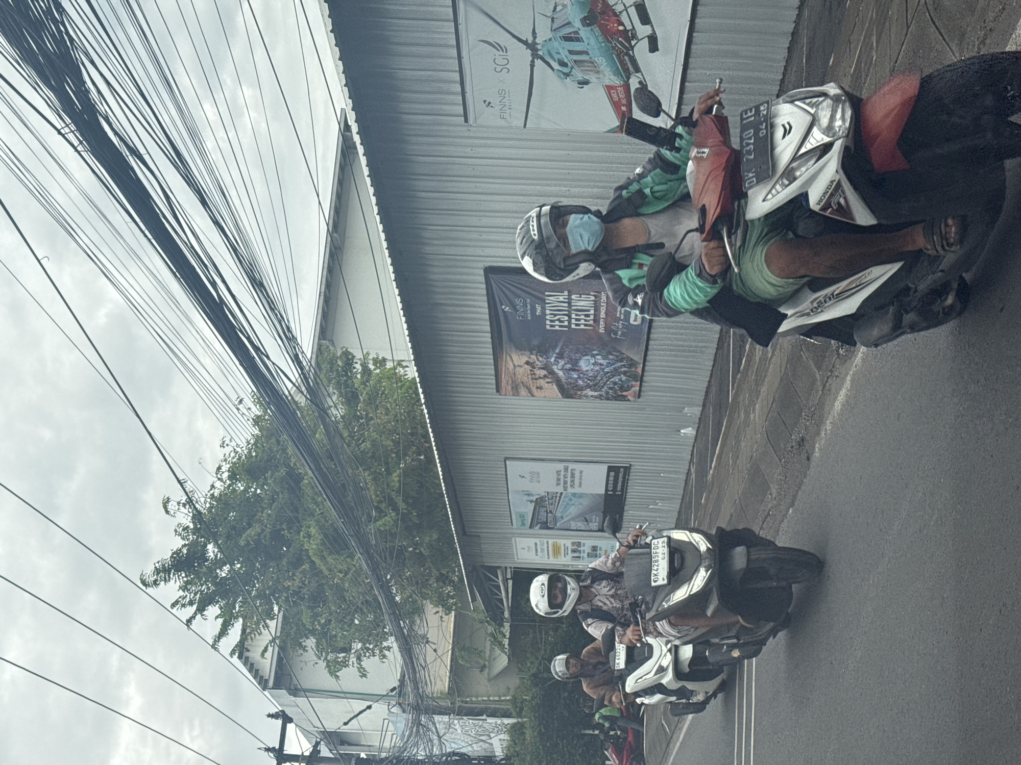 This image depicts a street scene in North Kuta, Indonesia, under an overcast sky. The perspective is slightly rotated, capturing a view across a paved road towards buildings and overhead utility lines.
In the midground, several individuals are riding motorbikes (scooters) on the road. The closest visible rider wears a light green jacket, a white full-face helmet, and a light blue face mask, operating a white and red Honda scooter. The license plate, "DK 2320 IE," is clearly visible, along with an expiry date of "04-28." Behind this rider, another person in a patterned shirt and a white helmet is also on a scooter, with their license plate displaying "DK 4289FDC 02-29." Other riders are partially visible further down the street, all wearing helmets.
To the right, behind the moving traffic, are buildings with corrugated metal facades. These structures display large advertising banners. One banner features "FINNS x SGI" with an image of a helicopter and the text "SEARCH AND RESCUE." Below it, another prominent banner reads "FINNS THAT FESTIVAL FEELING EVERY SINGLE DAY!" A third, partially visible banner mentions "FINNS THE ONLY HOTEL."
The upper section of the frame is filled with a dense tangle of black utility cables crisscrossing against the cloudy, light grey sky. To the left, a large, leafy green tree provides a natural element, its branches visible above the cables and buildings. The scene reflects a typical urban environment in Southeast Asia, characterized by scooter traffic and dense overhead wiring.