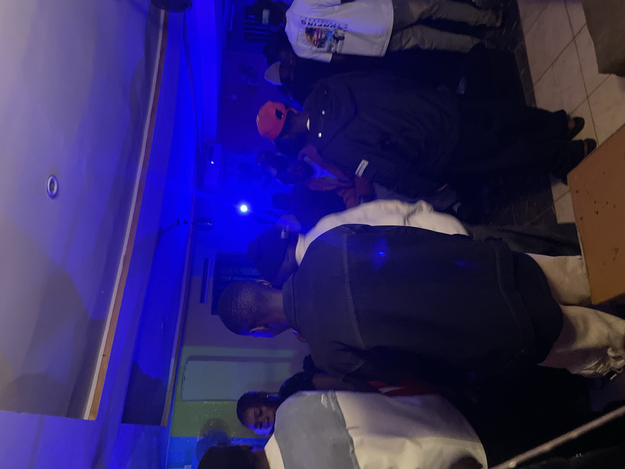 The image depicts an indoor gathering, likely a party or social event, taking place in Durumi, Nigeria. The lighting is predominantly blue, casting a dim and atmospheric glow throughout the space. Several individuals are visible, with their backs primarily turned to the camera, suggesting they are engaged with each other or a focal point not captured. The setting appears to be a venue with a tiled floor and a ceiling with integrated lighting fixtures. One individual in the foreground wears a dark jacket, another a light-colored jacket with a grey panel, and a third a white t-shirt with graphics. Another person is wearing a red cap. The overall impression is one of a lively social gathering.