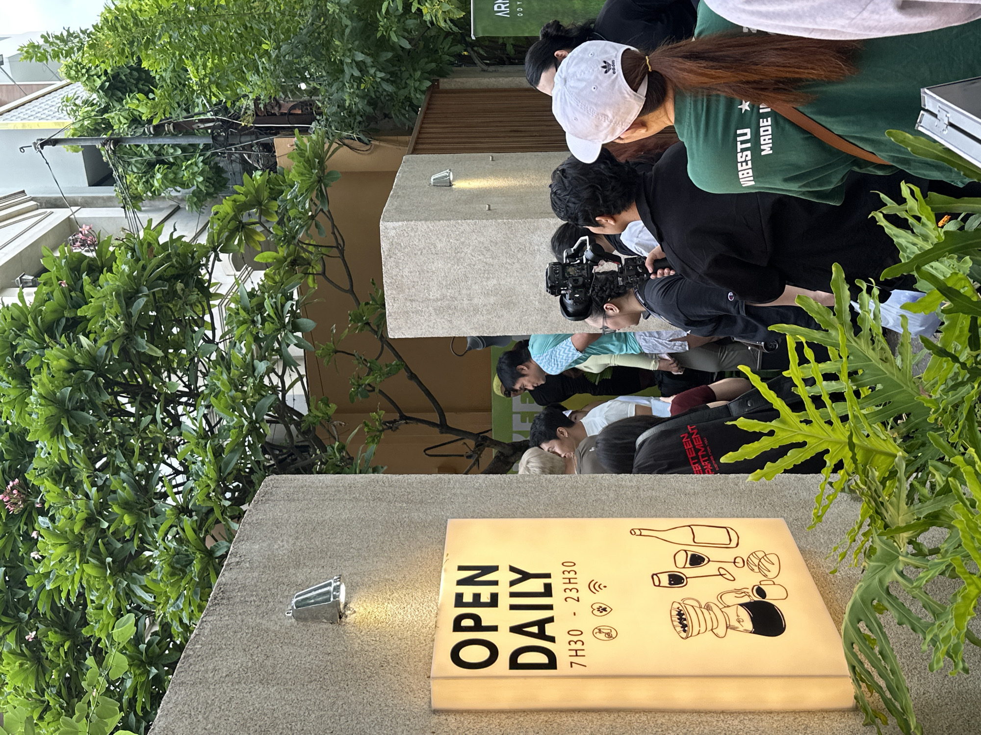 The image shows a group of people standing outside a building in Thủ Đức, Vietnam. A sign that reads "Open Daily 7H30 - 23H30" is visible on the building. A woman wearing a green shirt, an Adidas hat and a brown satchel is in the crowd. There is a person holding a professional video camera. There are various trees and plants throughout the frame.