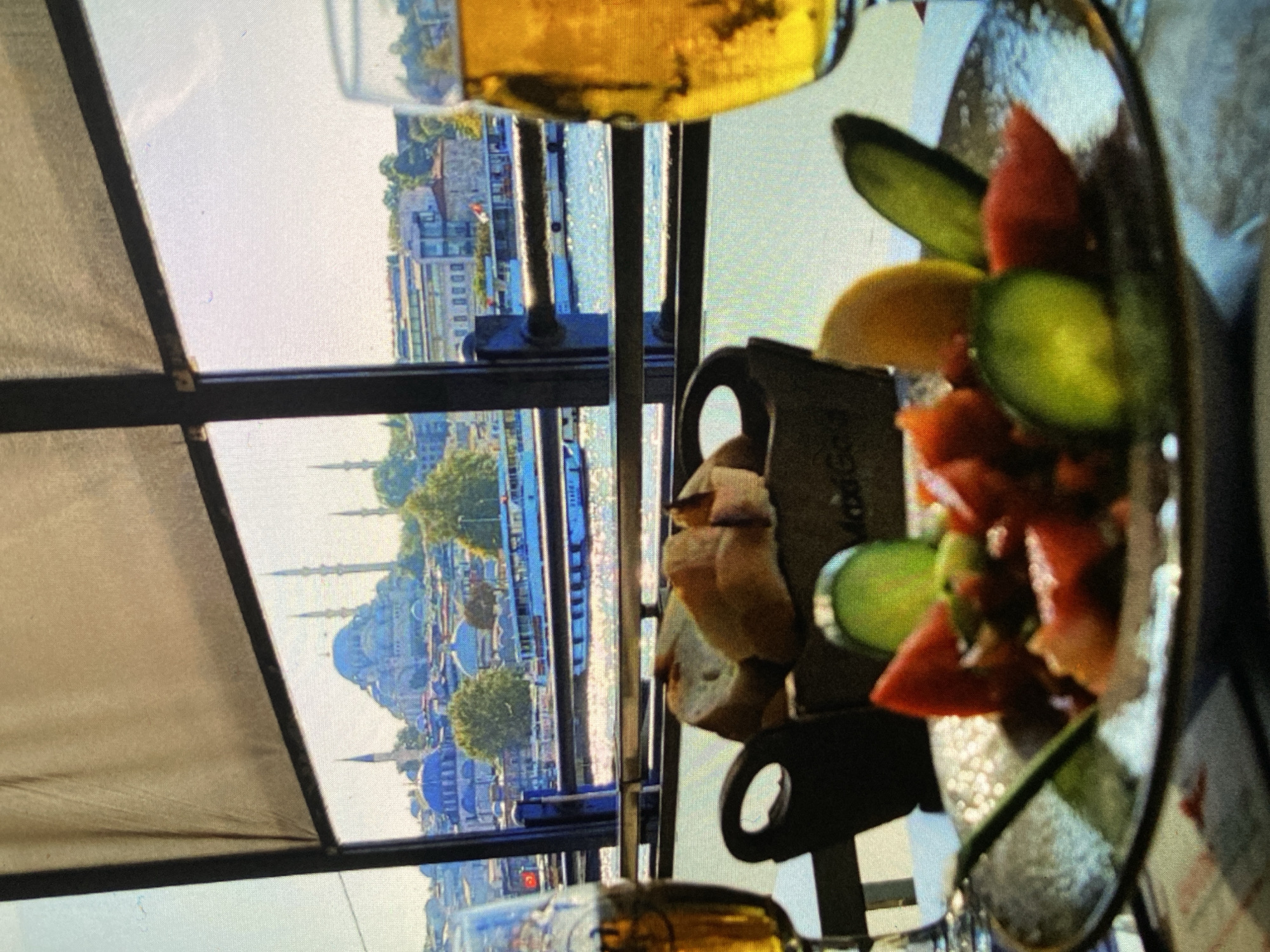 The image captures an outdoor dining experience with a panoramic city and water view, presented in a portrait orientation that, when viewed correctly, shows a horizontal vista. In the foreground, a glass of golden beverage (likely beer) is visible at the top, and another partially visible at the bottom. On a table, a plate features a colorful salad or appetizer with chopped red ingredients (possibly tomato or watermelon), green slices (cucumber), and yellow wedges (lemon or melon). Next to it, a dark, textured container holds pieces of bread or toast, with the word "MaxiGold" subtly visible on its side.
The setting appears to be a balcony or terrace, with dark metal railings and a light-colored canvas awning framing the expansive view. Beyond the railing, a wide body of water, possibly a strait or bay, is seen with boats. The background showcases a vibrant cityscape under a bright, clear daytime sky. The most prominent features are several large mosques with multiple domes and slender minarets, characteristic of Ottoman architecture, strongly suggesting the location is Istanbul, Turkey, rather than Bankya, Bulgaria. Trees and other buildings are scattered throughout the urban landscape.
The activity depicted is enjoying a meal or refreshments while taking in a scenic view. The bright lighting indicates it is daytime with good weather. The overall impression is one of relaxation and enjoyment.