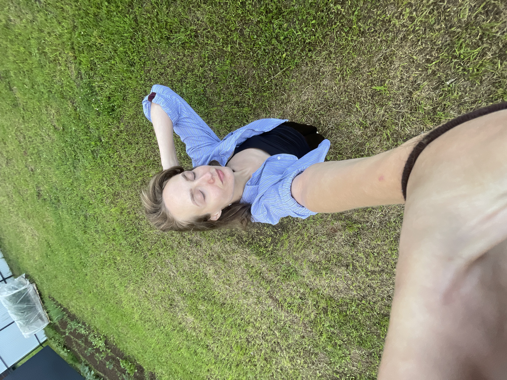 A young woman with light brown hair is lying on her back in a grassy field, taking a selfie. She is wearing a blue and white striped button-down shirt, unbuttoned and draped over a black top. Her eyes are closed, and her head is tilted back, suggesting a relaxed or peaceful state. One arm is bent with her hand resting behind her head, and the other arm is extended, holding the camera. The background is a lush green lawn, with some patches of drier grass. In the distance, on the left side of the frame, there is a small garden bed with rows of young plants, and a structure covered with a clear plastic sheet, possibly a small greenhouse or cold frame. The overall scene appears to be outdoors in a rural or suburban setting, likely during the daytime. The lighting suggests it could be late afternoon or early evening. The weather seems pleasant, conducive to outdoor activities. The emotional tone conveyed is one of tranquility and enjoyment of nature. No visible text is present in the image.
