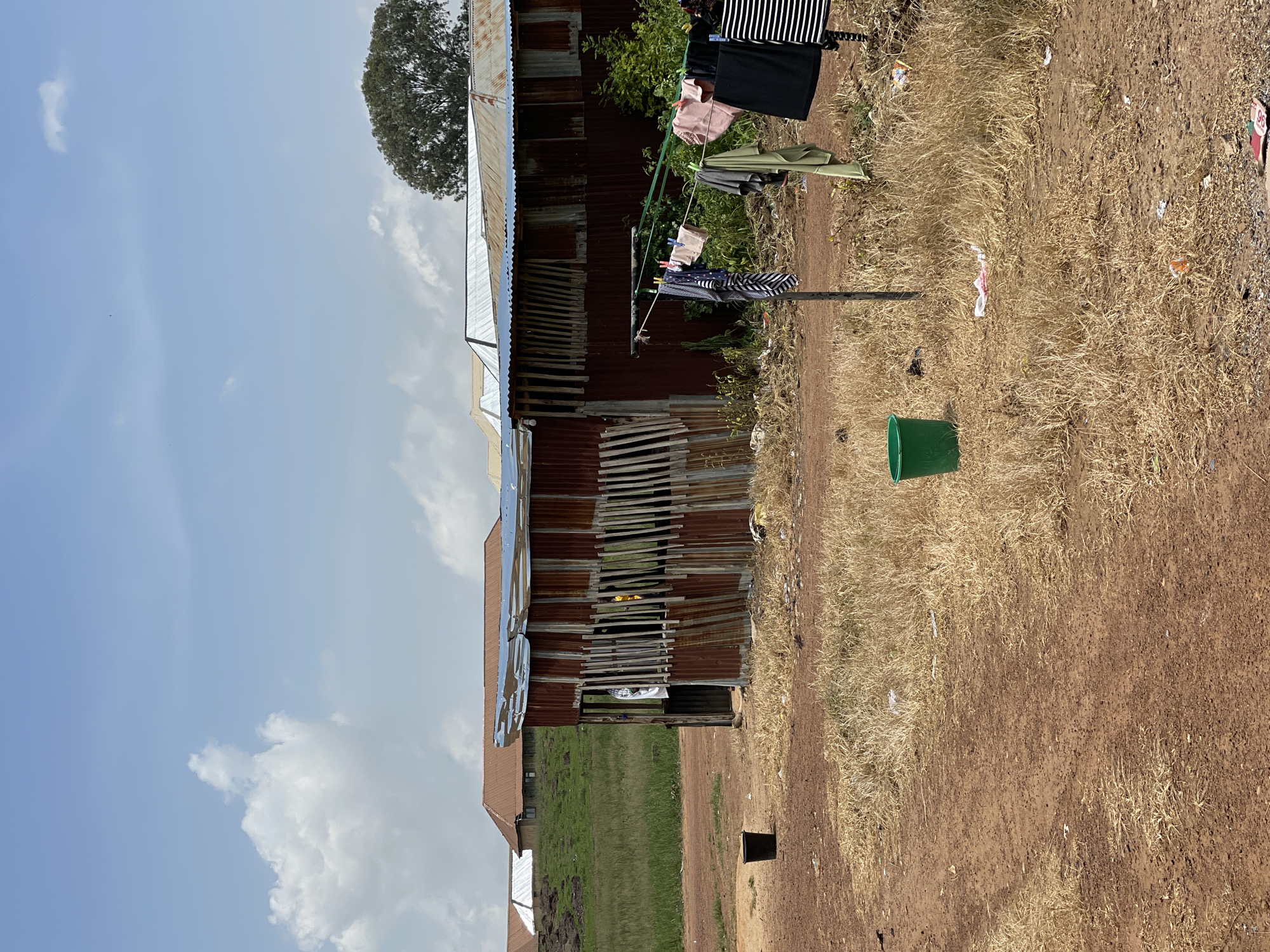 The image depicts an outdoor scene in Durumi, Nigeria, featuring a weathered corrugated metal structure that appears to be a makeshift dwelling or shop. The structure has a dilapidated roof and sides, with vertical wooden planks forming part of its facade. In front of it, laundry is hung on a clothesline, including various garments such as striped shirts, shorts, and dark-colored clothing. A green plastic bucket sits on the dry, brown earth to the left of the structure, and another green bucket is visible closer to the clothesline. The ground is covered in dry grass and scattered debris, suggesting a rural or semi-urban environment. In the background, several buildings with reddish-brown roofs are visible on a grassy hillside under a bright blue sky with scattered clouds. A large tree stands to the right of the background buildings.