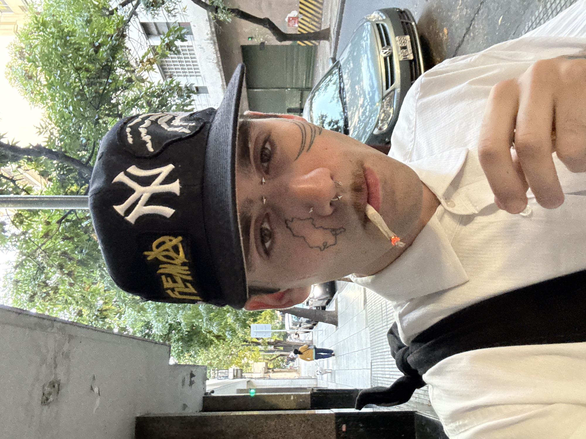 A fair-skinned young adult with multiple facial piercings and tattoos, wearing a black baseball cap with a white "NY" logo and a yellow inscription, is seen holding a lit cigarette in their mouth. The person is wearing a white shirt with a black stripe across the chest. The background includes a city street with a parked silver car, trees, buildings, and people walking in the distance. The location is likely in Buenos Aires, Argentina, based on architectural style and street features.
