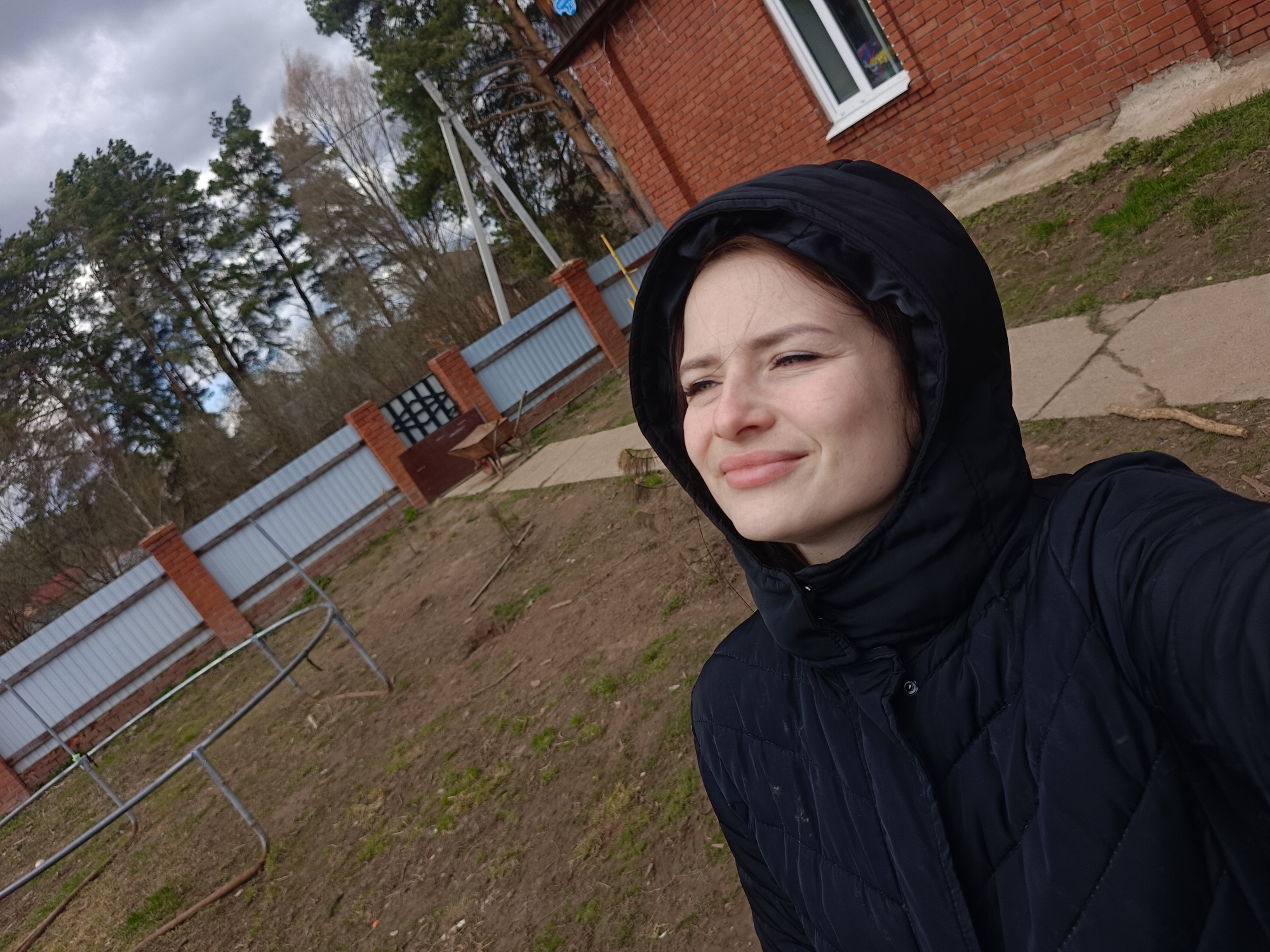 A young woman is featured in a close-up, selfie-style photograph, captured outdoors in Rusalevka, Russia. She has light brown hair partially visible beneath the hood of a black, quilted puffer jacket, which suggests cool weather. Her face is turned slightly to her left, away from the direct camera gaze, and she exhibits a subtle, pleasant smile, giving her a relaxed and contented expression.
The background depicts a semi-rural or suburban environment. To her right, a portion of a red brick house with a distinct white-framed window is visible. Further into the background, behind a sturdy fence, tall evergreen trees, likely pines, rise prominently against the sky. Some bare deciduous trees are also scattered among them, hinting at a late autumn or early spring season.
The fence features brick pillars interspersed with corrugated metal panels, and a dark-colored gate or section is visible on the left. A wheelbarrow rests behind this fence section. The ground in the foreground consists of bare soil mixed with patches of sparse, dry grass. A concrete path runs alongside the house. A metallic, curved framework structure lies on the ground in the mid-left, possibly part of a garden structure or play equipment. The sky overhead is heavily overcast with gray clouds, indicating a cloudy daytime. No discernible text is visible in the image.