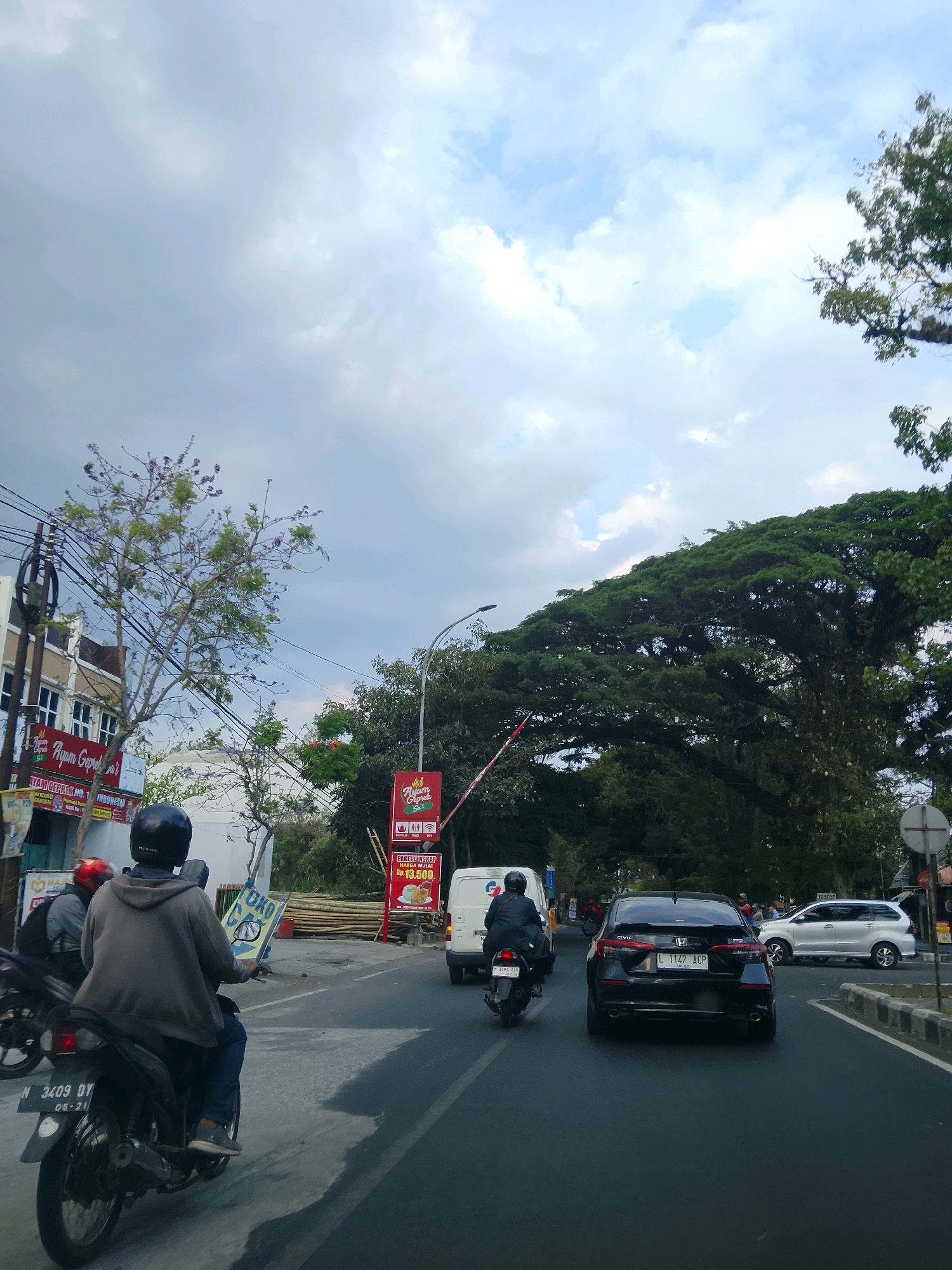 The image depicts a street scene in Malang, Indonesia, characterized by overcast skies and moderate traffic. In the foreground, a person is riding a black motorcycle, wearing a grey hoodie and a black helmet, with a red-helmeted passenger on the back. The motorcycle's license plate is visible as "N 3409 DT". Further down the road, a black Honda Civic with license plate "L 1122 ACP" is in motion, preceded by a white van. To the right, a silver SUV is parked on the side of the road.
The left side of the street features buildings with signage for "Ayam Geprek", indicating a food establishment. A jacaranda tree with purple flowers is in bloom near the buildings. The right side of the street is dominated by a large, mature tree with dense foliage, its branches extending over the road. Red and white signage, also advertising "Ayam Geprek", is visible near the large tree. Streetlights and power lines are present throughout the scene, typical of urban environments. The road surface appears to be asphalt, with visible lane markings. Bamboo stalks are stacked on the side of the road behind a barrier, suggesting ongoing construction or renovation. The overall atmosphere is that of a busy, everyday street in an Indonesian city.