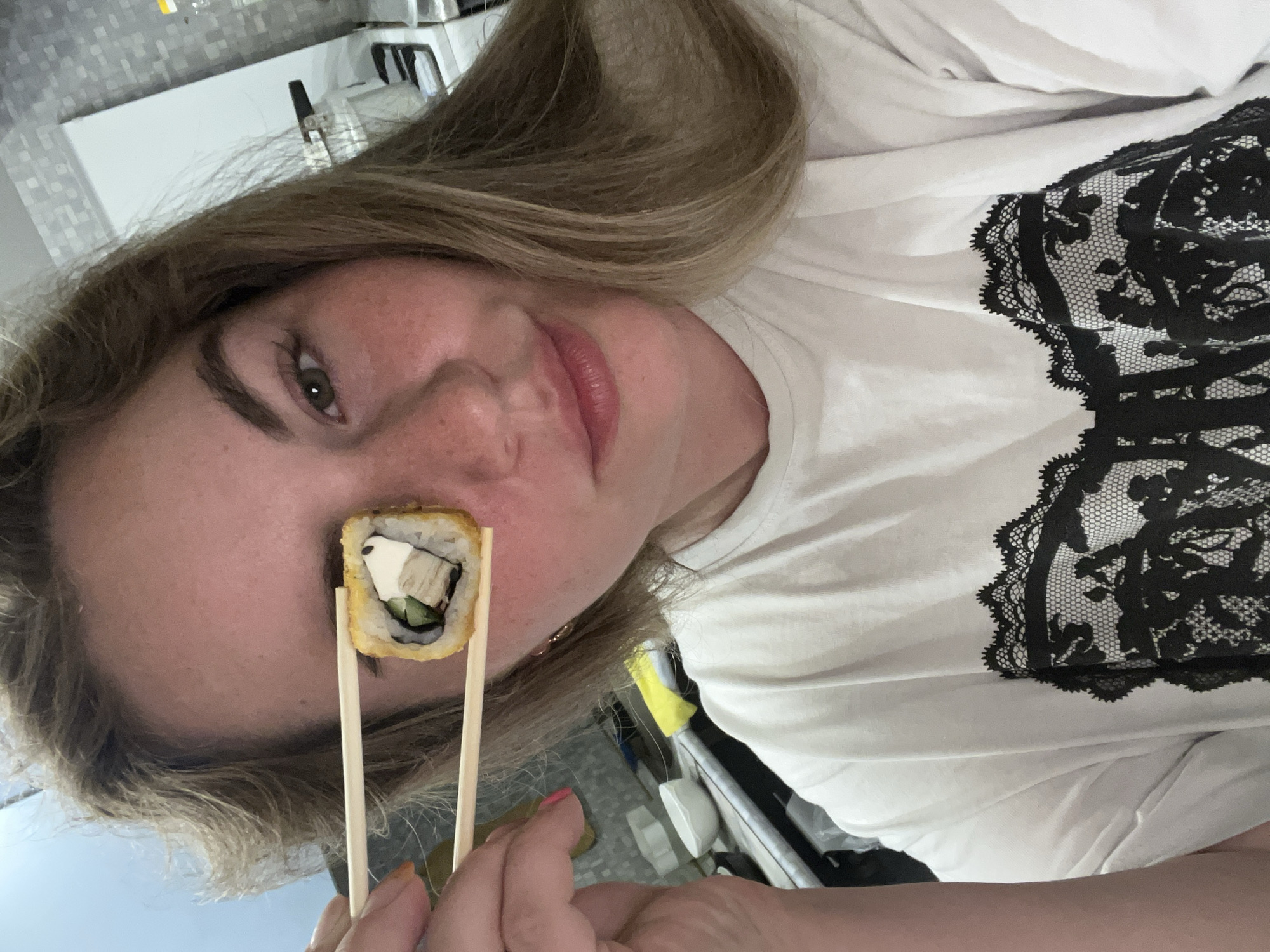 A woman is holding a piece of sushi with chopsticks up to her eye. She has blonde hair and fair skin. She is wearing a white shirt with a black lace design. The background appears to be a kitchen with white cabinets. There is not enough information to confirm the location in the city of Chaikovsky, Russia.