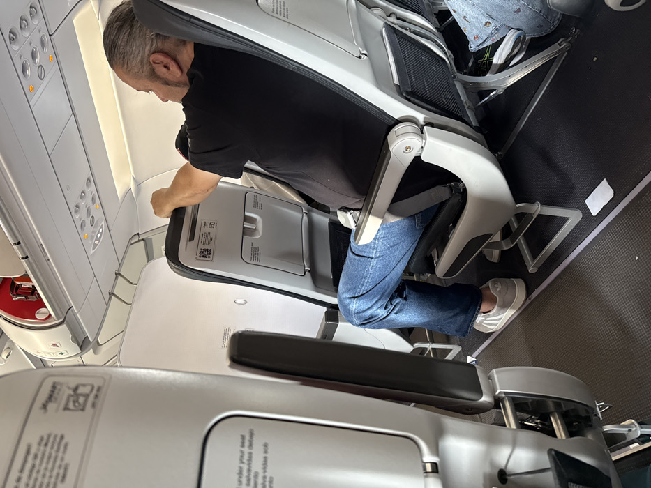 The image depicts the interior of a commercial aircraft cabin. The scene is reportedly situated within the context of AMCO, Area Metropolitana Centro Occidente, Colombia.
An adult male, with graying hair and a beard, occupies an aisle seat in the foreground-middle. He wears a black short-sleeved t-shirt, blue jeans, and light-colored athletic shoes with three dark stripes. His right arm is raised, with his hand resting on the top edge of the seatback directly in front of him. He is seated facing forward, leaning slightly. A gray seatbelt is visible across his lap.
The aircraft interior features white and light gray paneling, with dark gray/black seats. The seatbacks are equipped with tray tables and lower seat-back pockets. A "JetSMART" logo and instructional text in English ("under your seat") and Spanish ("salvavidas debajo") are visible on the seatback directly in front of the camera, to the left of the main subject. A QR code and additional text are visible on the seatback in front of the main subject.
Overhead, white storage compartments are present, one of which is partially open, revealing a red object inside. An overhead panel with circular light and vent controls is also visible. The floor is covered with a dark gray textured material, and a small, rectangular white object lies on the floor near the main subject's feet. Partial views of other passengers or their belongings are visible in the seats adjacent to and in front of the main subject.