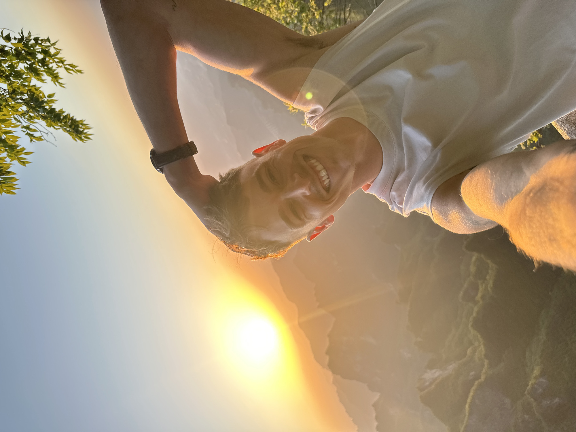 A low-angle, rotated, close-up shot shows a smiling Caucasian male in a white t-shirt, holding his left arm up, with a dark-colored watch visible on his wrist. In the background, a sunset or sunrise casts a warm glow over a mountainous, possibly forested, area. There are trees visible to the left and what appears to be a valley, presumably near Yongding District, China, given the prompt.