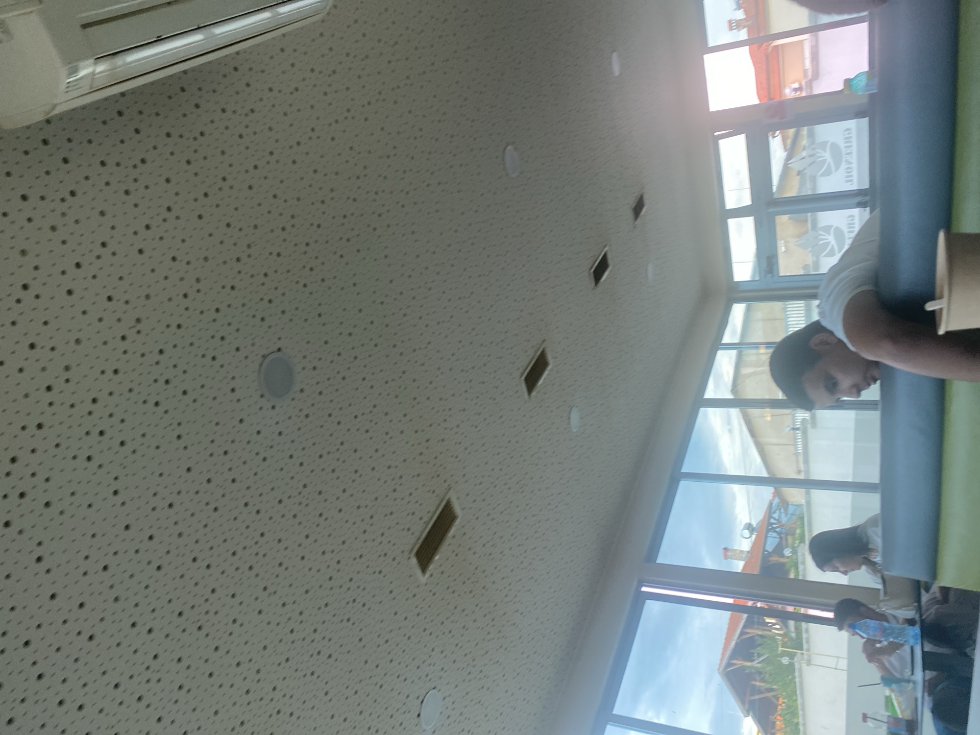 The image depicts an indoor scene, possibly a cafe or restaurant, with a focus on the perforated ceiling and the environment visible through large windows. The ceiling is white and covered in a dense pattern of small, dark dots. Several rectangular ventilation grates and circular light fixtures are embedded in the ceiling. To the right, part of a white air conditioning unit is visible.
Through the large windows, which span most of the left and middle sections of the image, a view of the exterior is presented. Buildings with red-tiled roofs and some wooden architectural elements are visible, suggesting a suburban or semi-rural setting. The sky is cloudy.
In the foreground, several people are seated. On the left, a person is partially visible, with a bottle of water and other items on a table in front of them. To their right, a woman is looking away, with dark hair. In the center-right, a young man in a white t-shirt is leaning back, appearing to be resting or sleeping. He is closest to the camera and his face is partially visible in profile. A disposable cup is in front of him on a cushioned bench, which is covered in green fabric. The bench extends across the bottom of the frame. The overall lighting suggests it is daytime, possibly late afternoon or early evening, given the quality of light visible through the windows. The location is identified as Ablanitsa, Bulgaria.