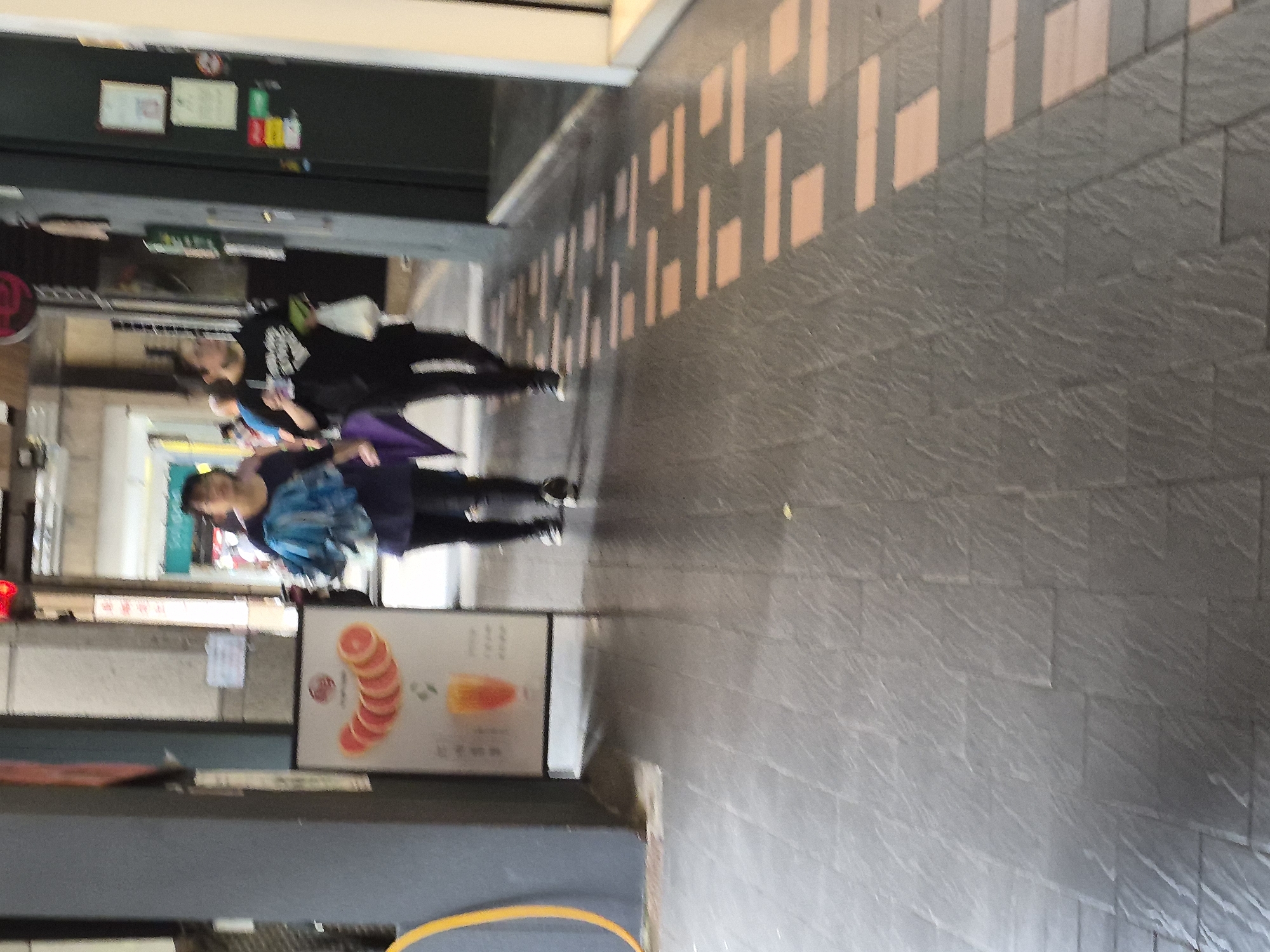 The image depicts a street scene, likely in an urban environment with a covered walkway. Two individuals are walking away from the camera, their faces not clearly visible due to the distance and resolution. The person on the left, wearing a dark top and pants, is carrying a blue umbrella or bag. The person on the right, also in dark clothing, holds a white plastic bag and a beverage cup.
In the foreground, the ground is paved with dark gray rectangular tiles. A sign is visible to the left, displaying an image of sliced grapefruit and a drink. Further down the walkway, the floor features a pattern of rectangular light-colored tiles interspersed with the darker ones. The background shows shop fronts and buildings, with some signage and lights. The overall impression is that of a commercial or pedestrian area. Based on the visual cues of the architecture and pavement, this scene is consistent with areas found in Taipei, Taiwan, known for its vibrant street life and covered walkways in commercial districts.