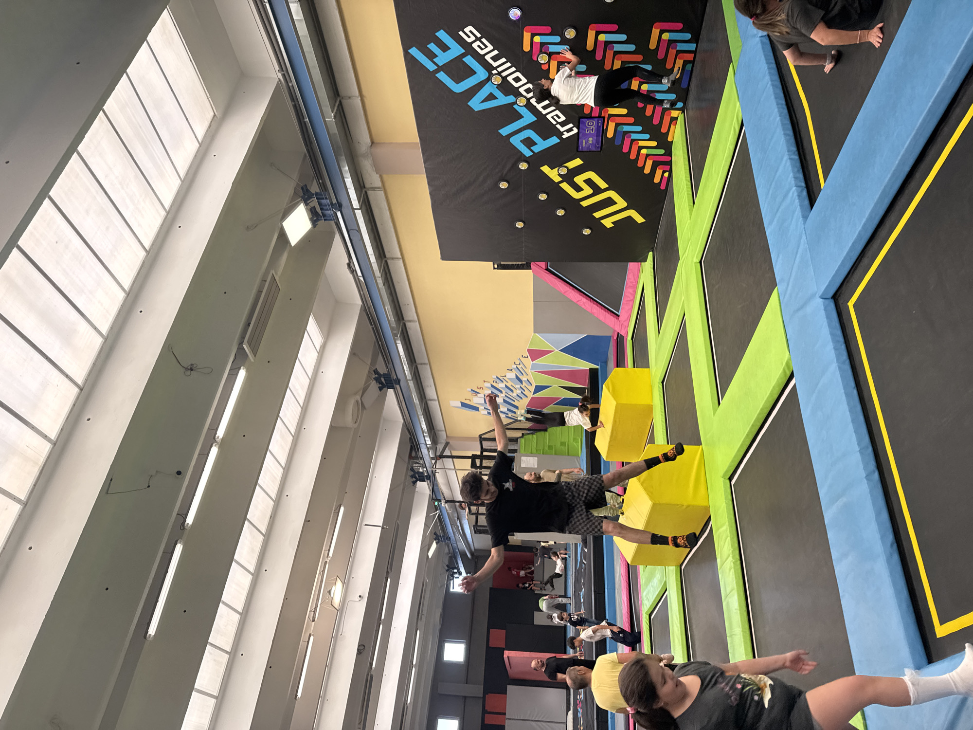 The image depicts the interior of "Just Place Trampolines," a trampoline park, likely located in Sofia, Bulgaria. The space is illuminated by overhead fluorescent lighting and natural light filtering through a row of high windows. There are multiple trampolines with colorful padding of blue, green, and yellow. Several people are visible, participating in activities on the trampolines. One individual is mid-jump with arms outstretched, wearing a black t-shirt and checkered shorts with black socks. The walls feature a branded design with the name of the establishment. Other individuals are either standing or moving around the trampoline area.