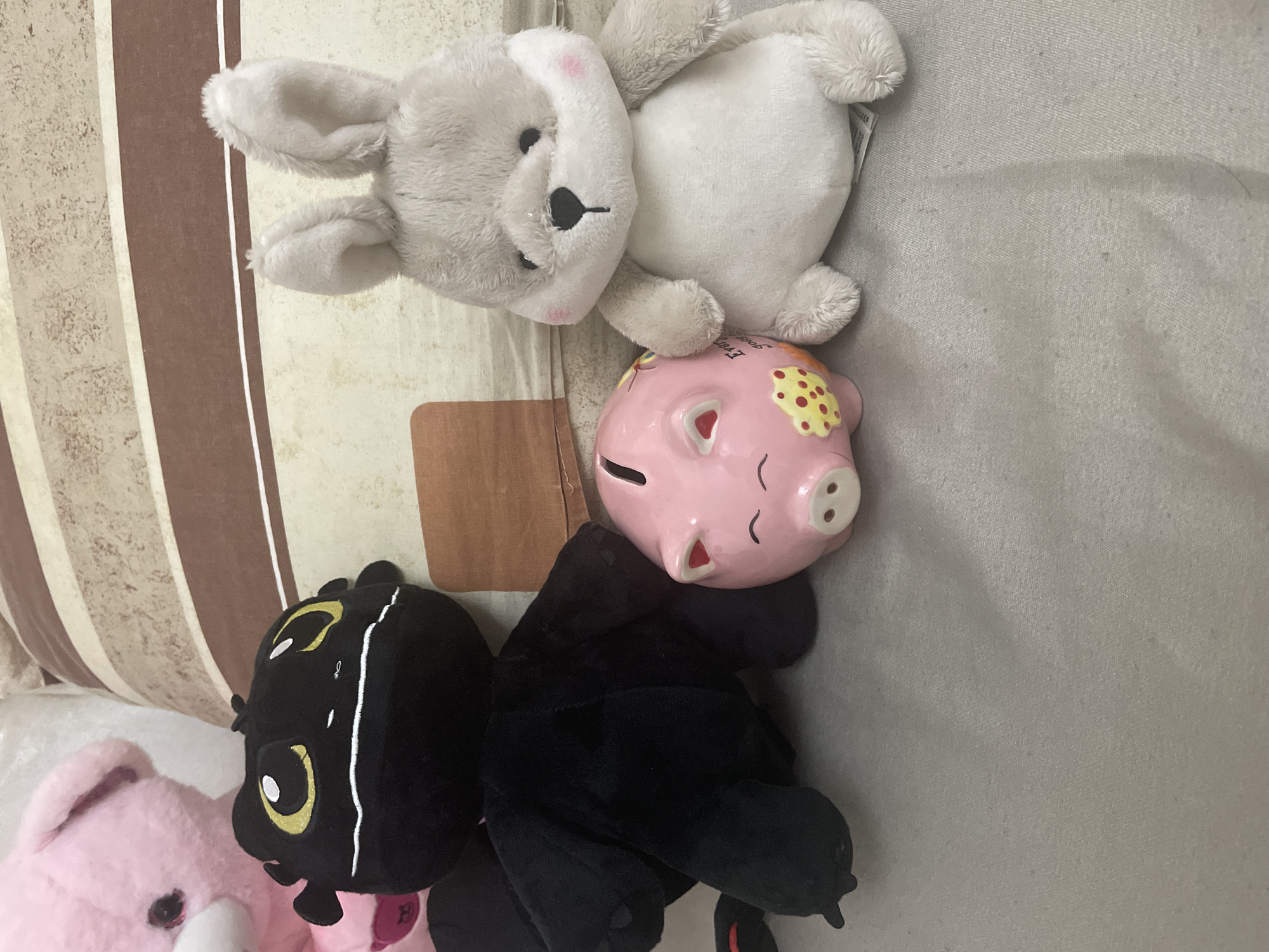 The image displays an indoor scene featuring three primary objects on a light grey fabric surface against a striped background.
From left to right, the objects are:
1. A black plush toy, resembling the character Toothless from "How to Train Your Dragon," positioned upright and facing slightly towards the right. It exhibits large yellow eyes with white pupils and a white stitched line across its snout. A portion of a pink plush toy is partially visible behind and to the upper left of the black plush toy.
2. A pink ceramic piggy bank, placed on its side between the black and grey plush toys. It features a smiling face with two small red-tipped ears and yellow polka-dot patterns on its visible side. A coin slot is present on its top surface.
3. A grey and white plush toy, identifiable as a rabbit or similar small mammal, positioned upright and facing forward. It has black bead eyes, a black stitched nose, and subtle pink blush marks on its cheeks. Its fur appears soft and textured.
All three main objects are situated on a light grey, possibly woven fabric surface, which occupies the lower half of the frame. The background consists of a striped fabric pattern, likely a cushion or pillow, featuring alternating horizontal bands of dark brown and a lighter, off-white or beige color. A small, rectangular, light brown object is partially visible resting on the striped background behind the black plush toy and piggy bank.
No individuals or explicit interactions are depicted. The environment suggests a domestic setting. The specific location, including the city of Ihtiman, Bulgaria, cannot be determined from the visual information presented in the image.