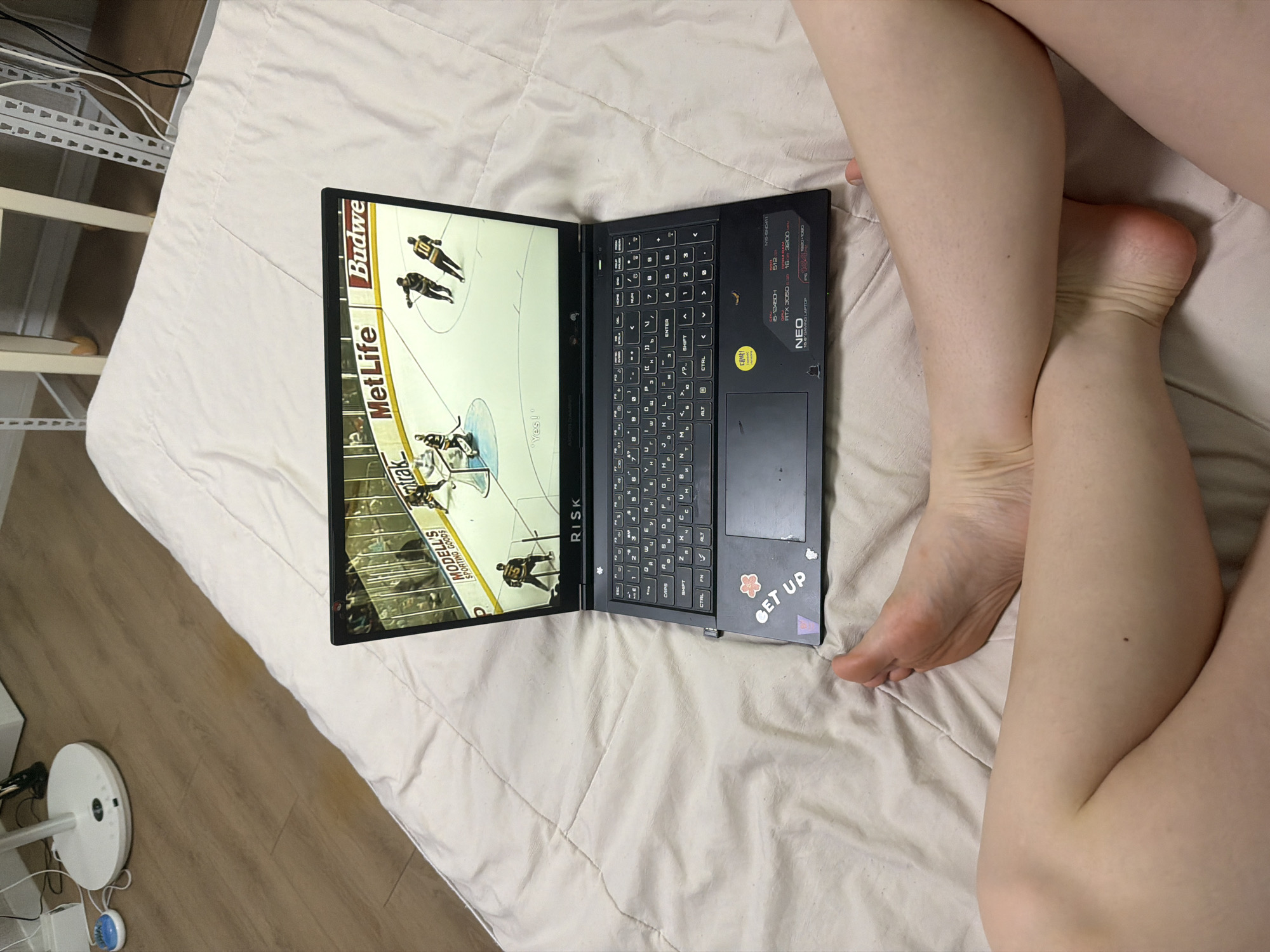A laptop is open on a bed, displaying a hockey game. The laptop has stickers on its surface, including one that says "GET UP." To the right of the laptop, two legs and feet are visible, positioned on the bed. The background shows a wooden floor and a white piece of furniture. The location is Dongducheon-si, South Korea.
