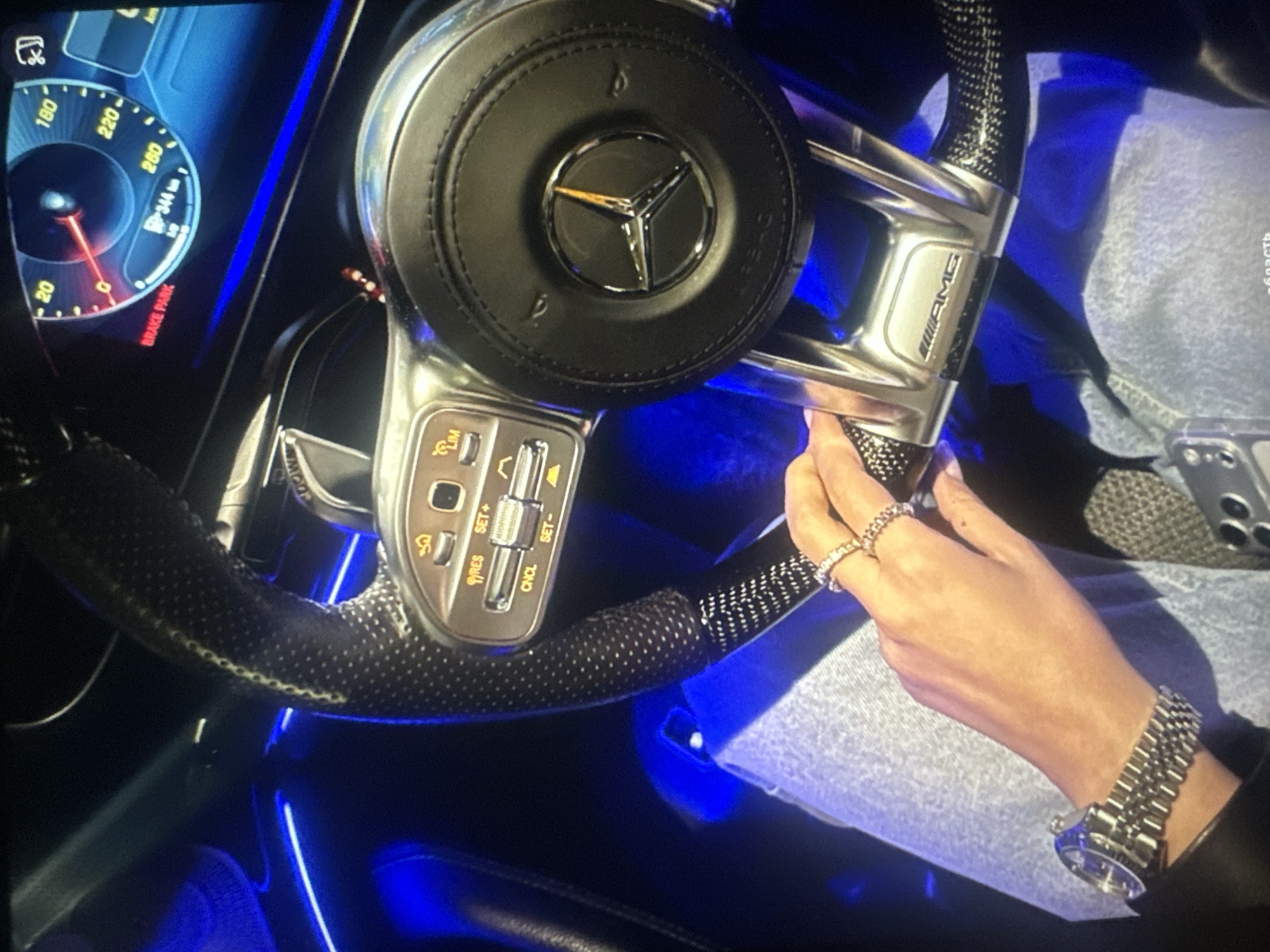 The image shows a close-up view of the interior of a Mercedes-Benz vehicle at night, illuminated by blue ambient lighting. The focus is on the steering wheel and the driver's hand. The steering wheel is made of black leather with carbon fiber accents and features the Mercedes-Benz logo in the center. The dashboard displays a digital instrument cluster with a speedometer and fuel gauge. A hand with two diamond rings and a silver wristwatch is resting on the steering wheel. The driver is wearing light-wash denim jeans. The background is dark and blurred, with hints of blue light reflecting off surfaces.