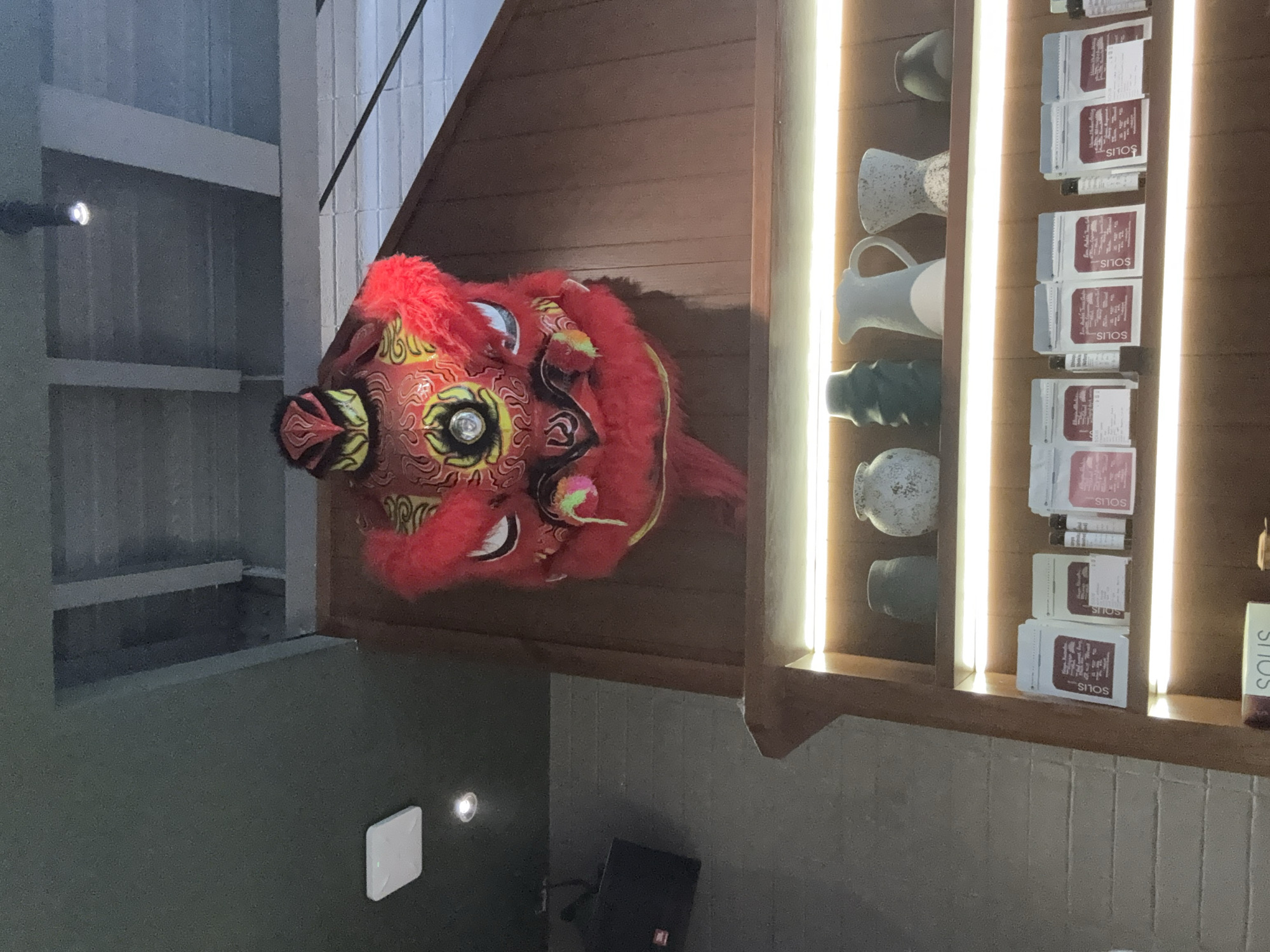 A red lion dance head sits atop a cabinet. The cabinet includes decorative ceramics and packages with the word "SOLIS" printed on them. The scene is inside a building. The location is Đà Nẵng, Vietnam, but this cannot be confirmed from the visual information.