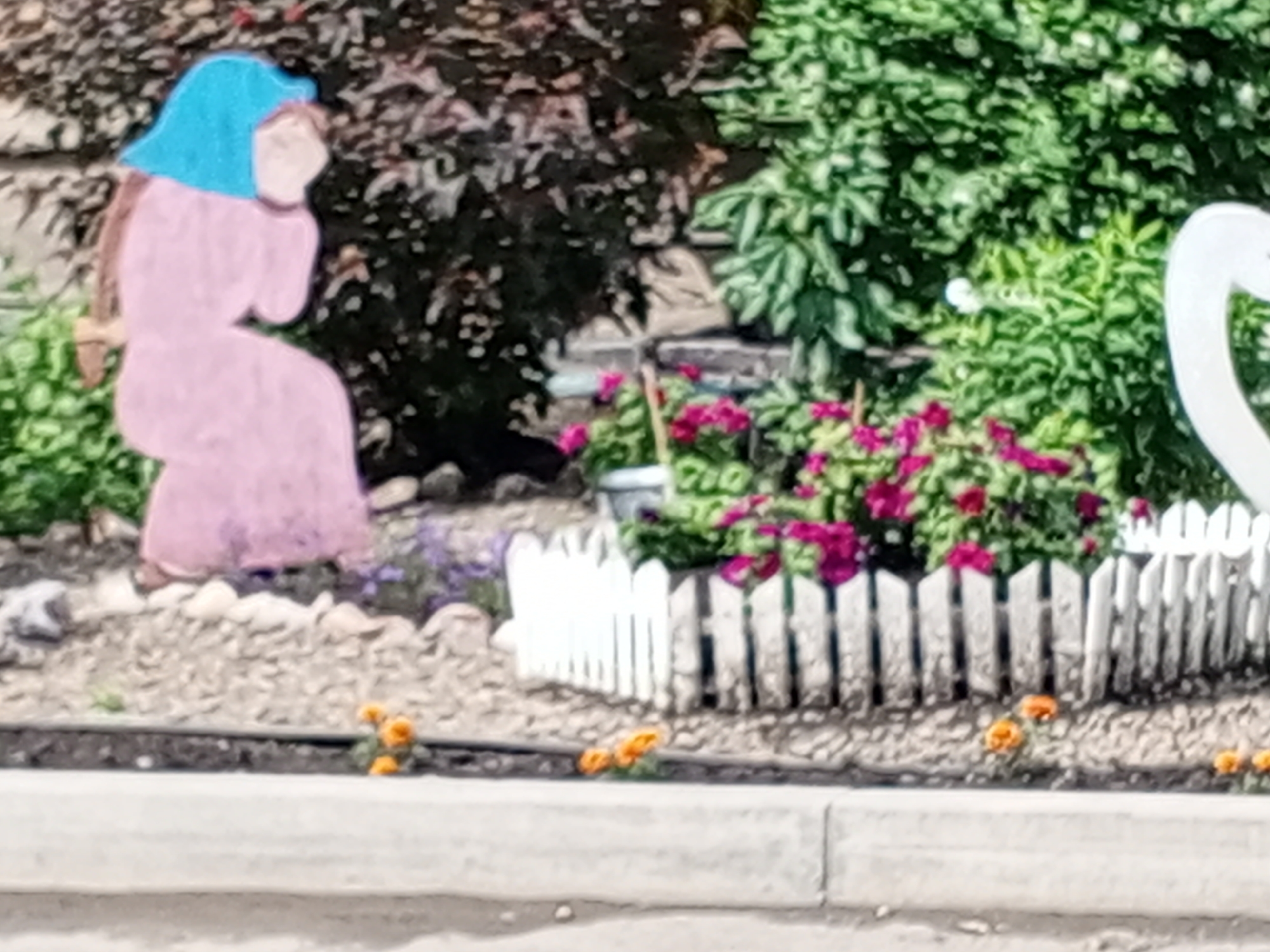 The image depicts a garden scene. In the foreground, a miniature white picket fence separates a gravel bed from a sidewalk or border. Several small orange flowers are planted in the gravel bed. Behind the fence, there is a planter with vibrant pink flowers. To the left, a flat, cut-out figure of a person, likely made of wood or similar material, is positioned. This figure is painted in shades of pink and blue and appears to be seated or kneeling. In the background, there are lush green bushes and darker foliage, with some hints of stone or brickwork. The overall impression is of a decorative garden display. The specific location in Krasnoyarsk, Russia, cannot be determined from the visual information provided in the image.