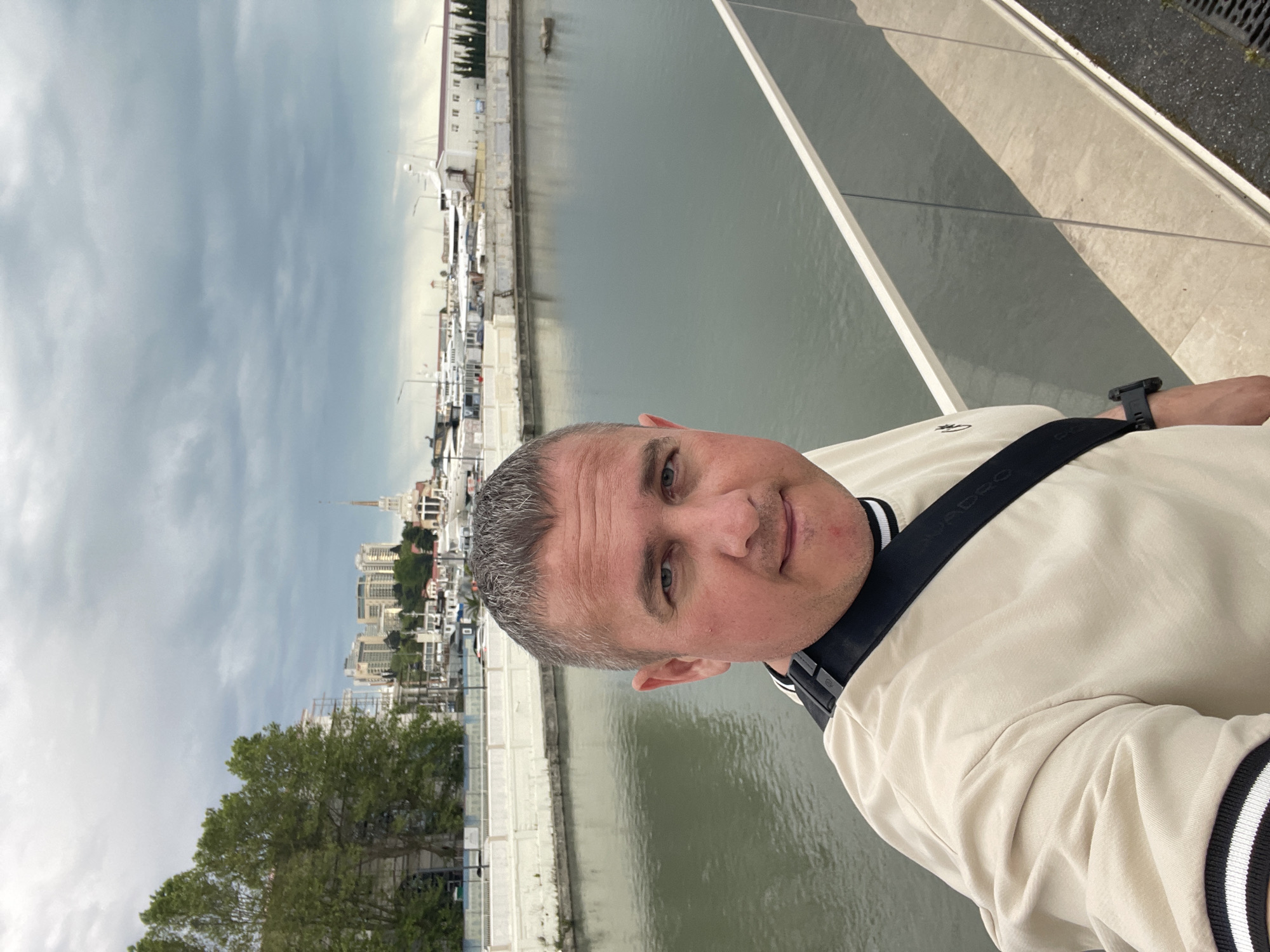 A man is seen in a medium close-up selfie shot. He is fair-skinned with salt and pepper hair. He is wearing a light beige jacket with black and white striped trim on the sleeves and a black shoulder strap. He is wearing a dark colored watch on his left wrist.
In the background is a waterfront cityscape. There is a harbor with many boats. Buildings are present as well as a tower or spire in the center of the cityscape. To the left, there is a green tree. The sky is overcast.
Based on the cityscape architecture, the image is likely taken in Sochi, Russia.