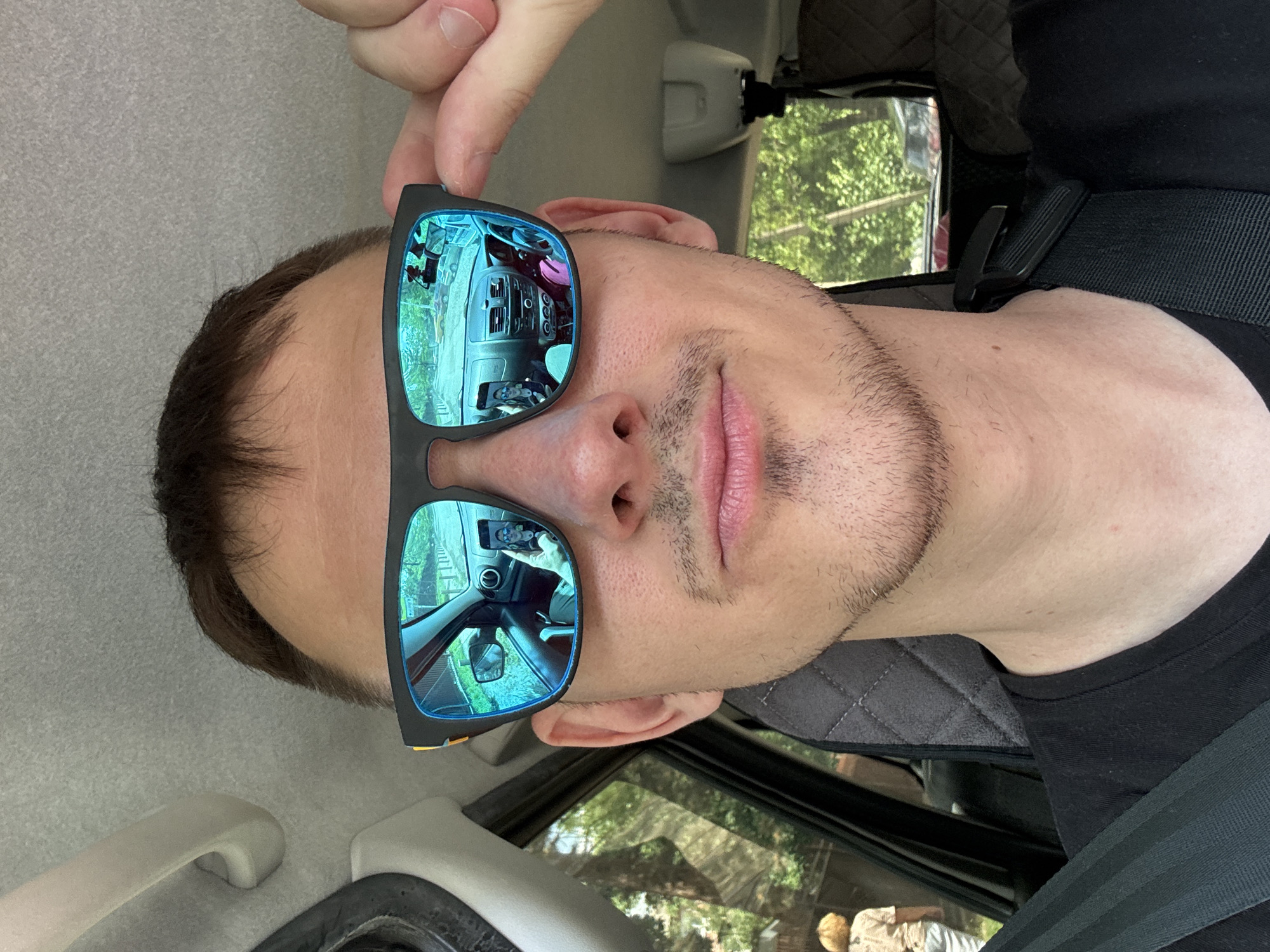 A man with sunglasses on, is in a car, secured by a seat belt. The reflection in the sunglasses reveals the car's dashboard and steering wheel. The environment appears to be outdoors with trees visible through the car windows. It is not possible to determine the specific location within Krasnodar, Russia from the image.