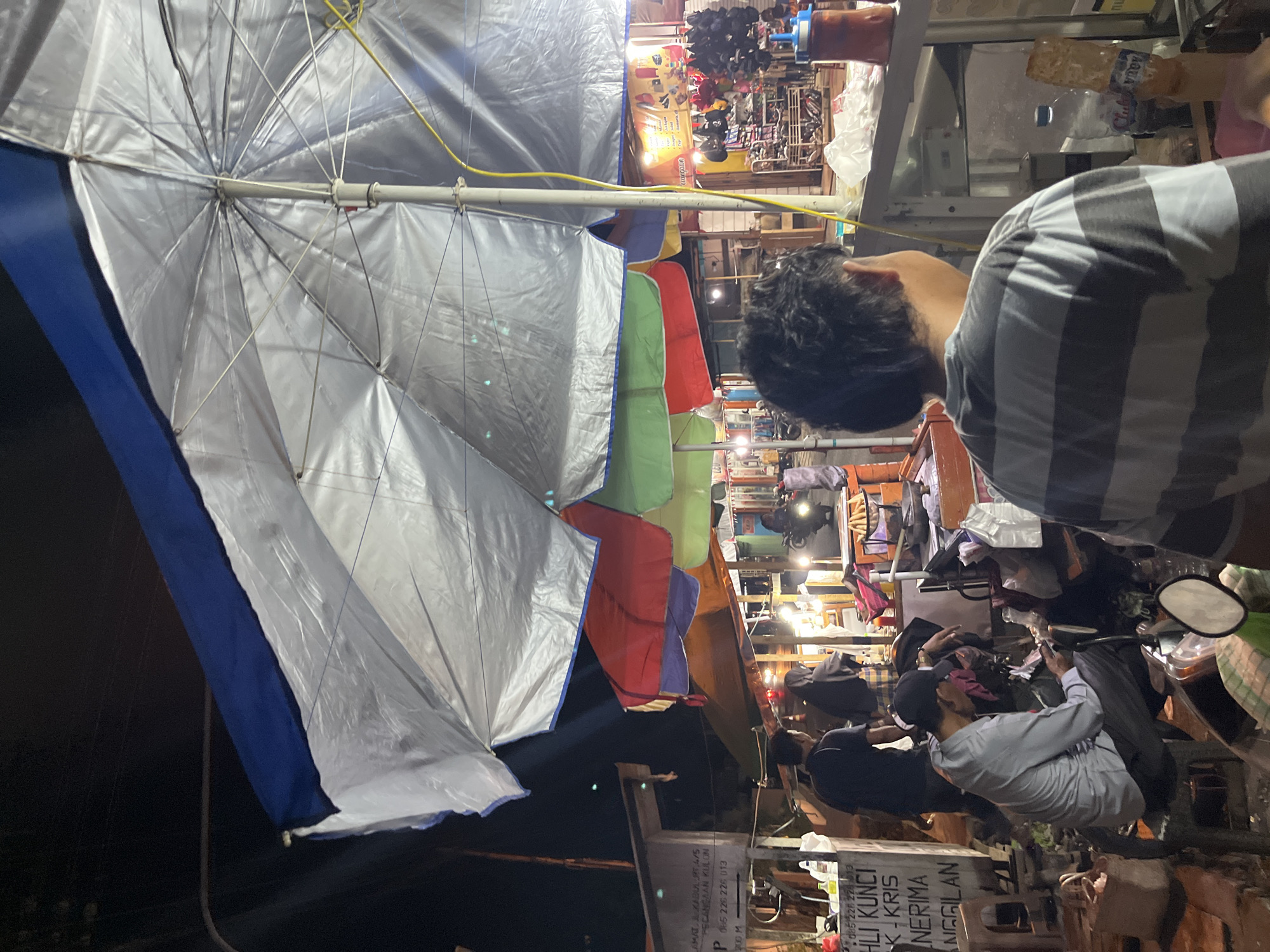 The image depicts a nighttime scene at a street food stall in Pecangaan, Indonesia. A large, silver umbrella with a blue trim is prominently featured, casting a reflective sheen. Beneath it, several other colorful umbrellas—red, green, and blue—overlap. Several people are visible in the foreground and midground, some appearing to be customers and others vendors. One man in a grey shirt and black cap is seated on a stool, looking at his phone. Another person, seen from the back, wears a striped t-shirt. Behind them, a busy market stall displays various goods, including hats and what appear to be food items. A motorcycle is also visible on the right side of the frame. The overall environment suggests a bustling night market or roadside food area.