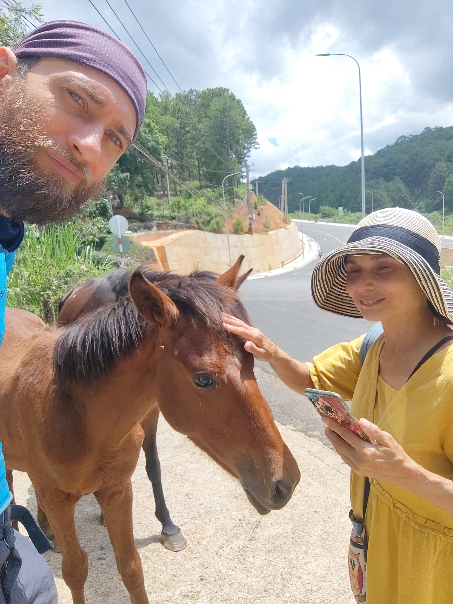 A selfie shows two people and a horse standing on the side of a paved road. The man on the left has a beard and is wearing a purple headband. The woman on the right is wearing a large straw hat and a yellow dress, holding a cell phone, and petting the horse's forehead with her right hand. The horse is brown with a dark mane. The road curves around a small hill in the background. In the distance, there are trees and a cloudy sky. The setting is likely in the outskirts or a rural area of Da Lat, Vietnam, as indicated by the local vegetation and the general road infrastructure.