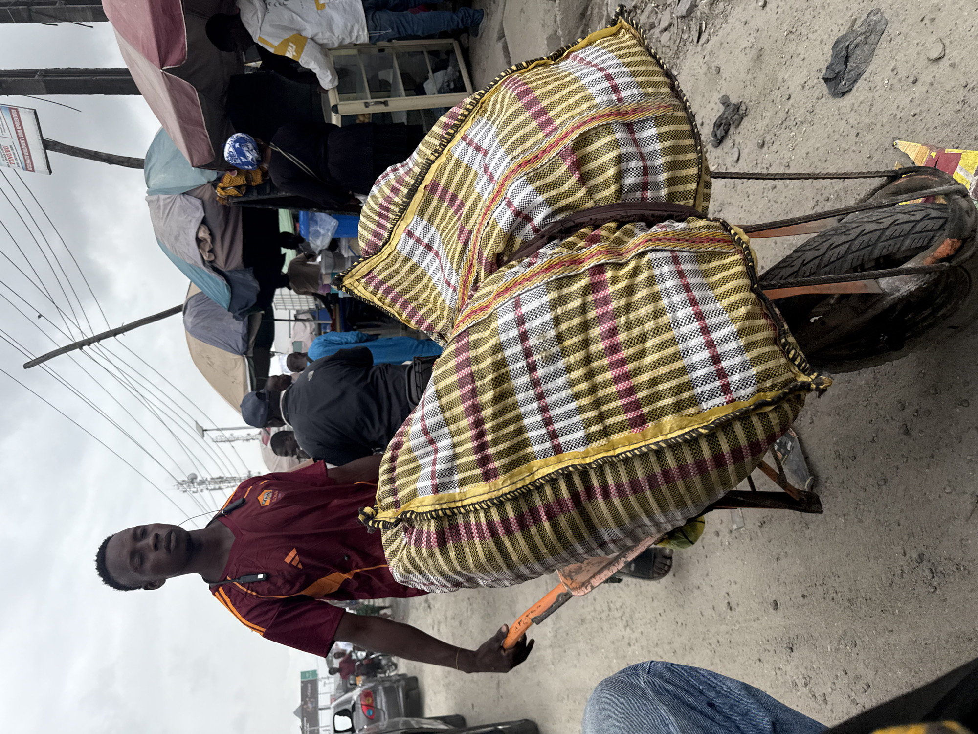The image depicts a street scene in Lekki, Nigeria, under an overcast sky. In the foreground, a young Black man is pushing a wheelbarrow loaded with two large, plaid-patterned bags. He is wearing a maroon and orange V-neck t-shirt with athletic stripes and has earbuds around his neck. The wheelbarrow has a metal frame and a single tire.
Behind the man and the wheelbarrow, a busy street unfolds. Several market stalls with colorful awnings are visible, selling various goods. People are milling around, some interacting with vendors, while others appear to be on motorcycles or in vehicles. Cars are present on the road, suggesting traffic congestion. The ground is a mix of asphalt and loose gravel. Overhead, power lines and communication towers are visible, indicating an urban environment. The overall impression is one of a bustling market or street commerce in a Nigerian city.