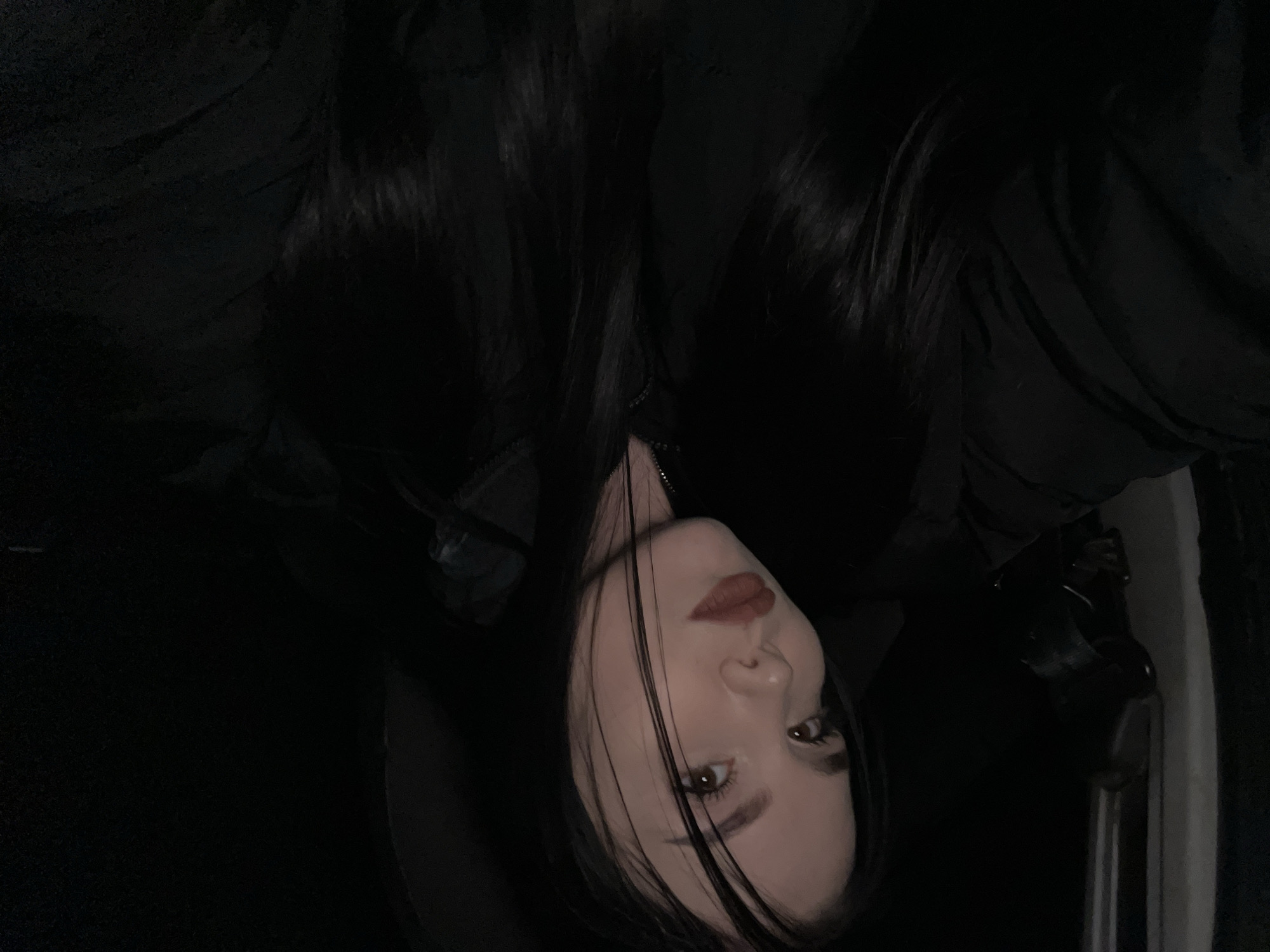 This is a dimly lit, close-up portrait of a young woman, captured from an elevated and tilted perspective. Her head appears angled in the frame, with long, straight, dark hair falling across her face. She has dark eyebrows and brown eyes, and her lips are painted with reddish-brown lipstick. Her skin tone is fair. She is wearing dark attire, possibly a jacket, with a zipper subtly visible near her neckline.
The woman's expression is neutral and direct, as she looks towards the camera. The background is exceptionally dark, providing minimal environmental context, appearing almost entirely black. Subtle textures within the darkness might suggest fabric, such as clothing or an interior lining of a vehicle or room. There is no discernible activity taking place other than the posing for the portrait.
The image conveys a sense of night or a very dimly lit indoor setting due to the lack of ambient light. No specific weather conditions are visible, and there are no clear location cues within the image itself. No text is visible. This photo originates from Chaikovsky, Russia.