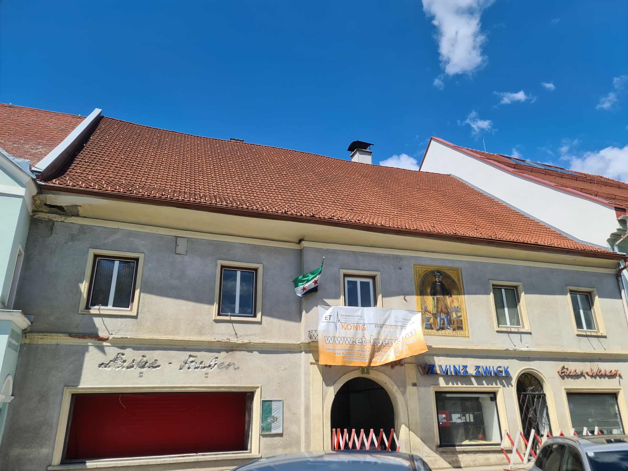 The image shows a building facade under a blue sky with scattered clouds in Sankt Marein bei Neumarkt, Austria. The building has a reddish-brown tiled roof and a light-colored stucco exterior. There are several windows on the upper floor, some with white frames and others with darker frames. A Syrian flag is hanging from one of the upper windows.
Below the windows, there is a large banner with text and a logo, reading "ET KÖNIG" and "www.et-koenig.at". The banner is orange and white. Further down on the facade, there are two shopfronts. The left shopfront has a large red covering or awning. The right shopfront has signage that appears to read "VZ VINZ. ZWICK" and "Eisen Waren". In front of the shops, there are barriers made of red and white striped plastic.
A fresco or mural depicting a man in historical attire holding a hammer and anvil is painted on the upper part of the facade to the right of the Syrian flag. There are also some inscriptions on the stucco above the shopfronts. The overall impression is of a commercial building in a town.