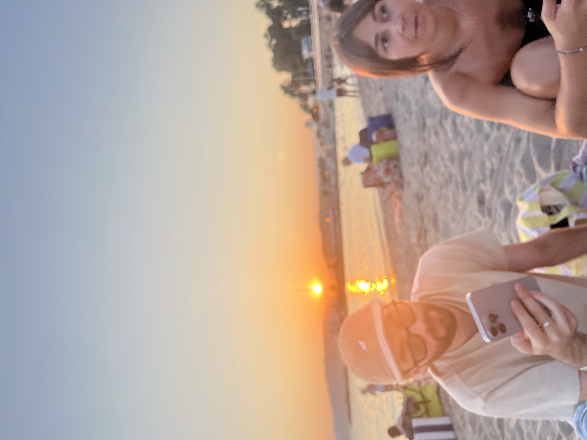 The image shows a beach scene during sunset in Cannes, France. The sun is low on the horizon, casting a warm glow over the water and the beach. In the foreground, a man wearing a cap and sunglasses is looking at his phone, with a ring on his left hand. To his right, a woman with short brown hair is partially visible, looking to the side. Various beach bags and towels are scattered on the sand. In the background, other people are visible on the beach and in the water, with some buildings and palm trees along the coastline. The overall atmosphere is relaxed and serene.