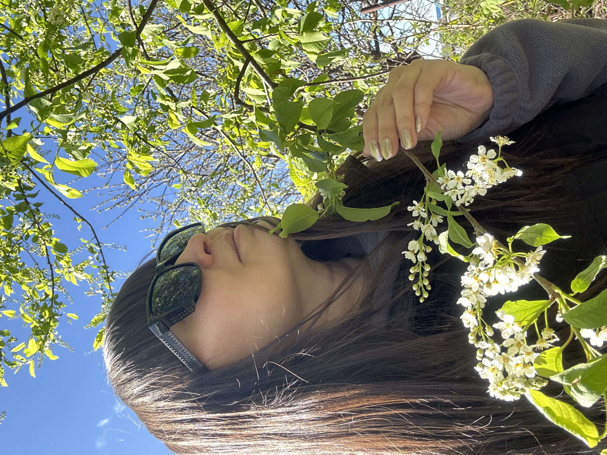 A woman with long, dark hair is seen from a low angle, looking up into a tree. She is wearing sunglasses. Her right hand is near a branch of white flowers and green leaves. The sky is blue with sparse clouds visible through the foliage.
