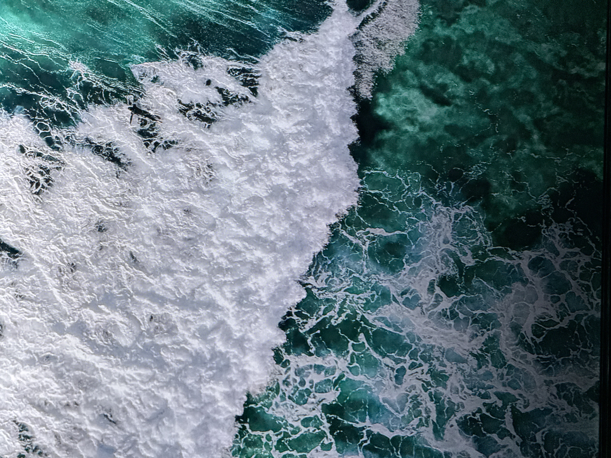 An aerial, extreme close-up shot captures the dynamic motion of crashing waves. The top portion of the image is dominated by a thick, white crest of foam, with intricate patterns and textures. This foam transitions downwards into the deep, turquoise-green waters of the ocean, where more subtle white foam swirls and churns, creating a mesmerizing, abstract pattern. A single dark silhouette of a surfer can be seen on their board in the upper right quadrant, positioned on a wave that is just beginning to break. The water is turbulent, with various shades of green and blue indicating depth and light refraction. The scene evokes a sense of power and natural beauty.