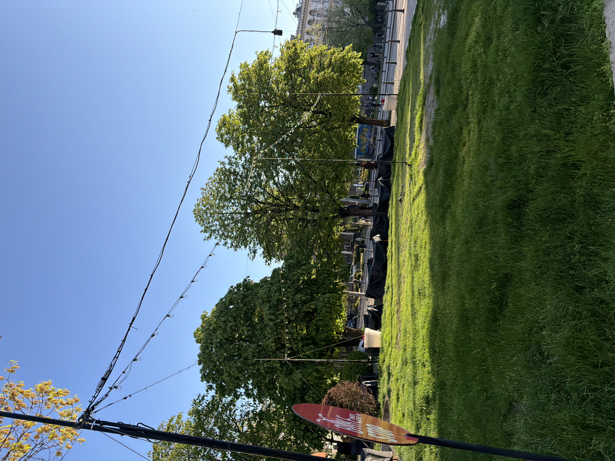 The image captures a scene in what appears to be a park in Sofia, Bulgaria. The view is oriented vertically, with the ground extending across the right side and the blue sky dominating the upper portion. In the foreground on the right is a grassy area. In the middle distance, there are trees, and covered tables suggesting a setup for an event. A signpost with a red sign is also visible in the lower portion. Overhead, a line of string lights spans across the scene. In the background, buildings typical of Sofia can be seen.