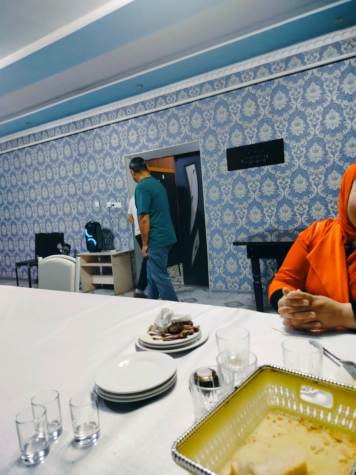 In the city of Көлкент, Kazakhstan, a dimly lit interior scene depicts a dining table set for a meal. Two men, one wearing a teal t-shirt and blue jeans, and another in a white shirt, are walking away from the camera towards a doorway. A woman, partially visible on the right side of the frame, wears an orange hijab and jacket. She is seated at the table, her hands clasped. The table is covered with a white tablecloth, on which plates of food, empty glasses, and a yellow basket containing bread are arranged. The room features patterned blue wallpaper and a white ceiling with blue accents. A black speaker with illuminated lights is positioned on a small table near the doorway.