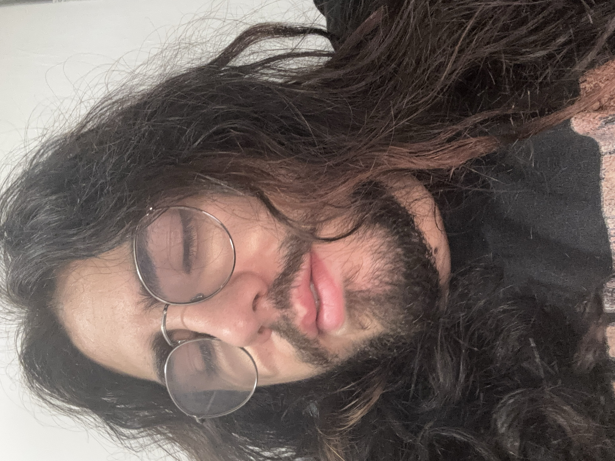 A close-up, low-angle, medium shot captures a person with dark, long, wavy hair and a beard, wearing round, wire-rimmed glasses. Their eyes are closed, and their lips are slightly parted. The lighting is soft and diffused, creating subtle shadows. The background is a plain, light-colored wall. The image appears to be taken indoors.