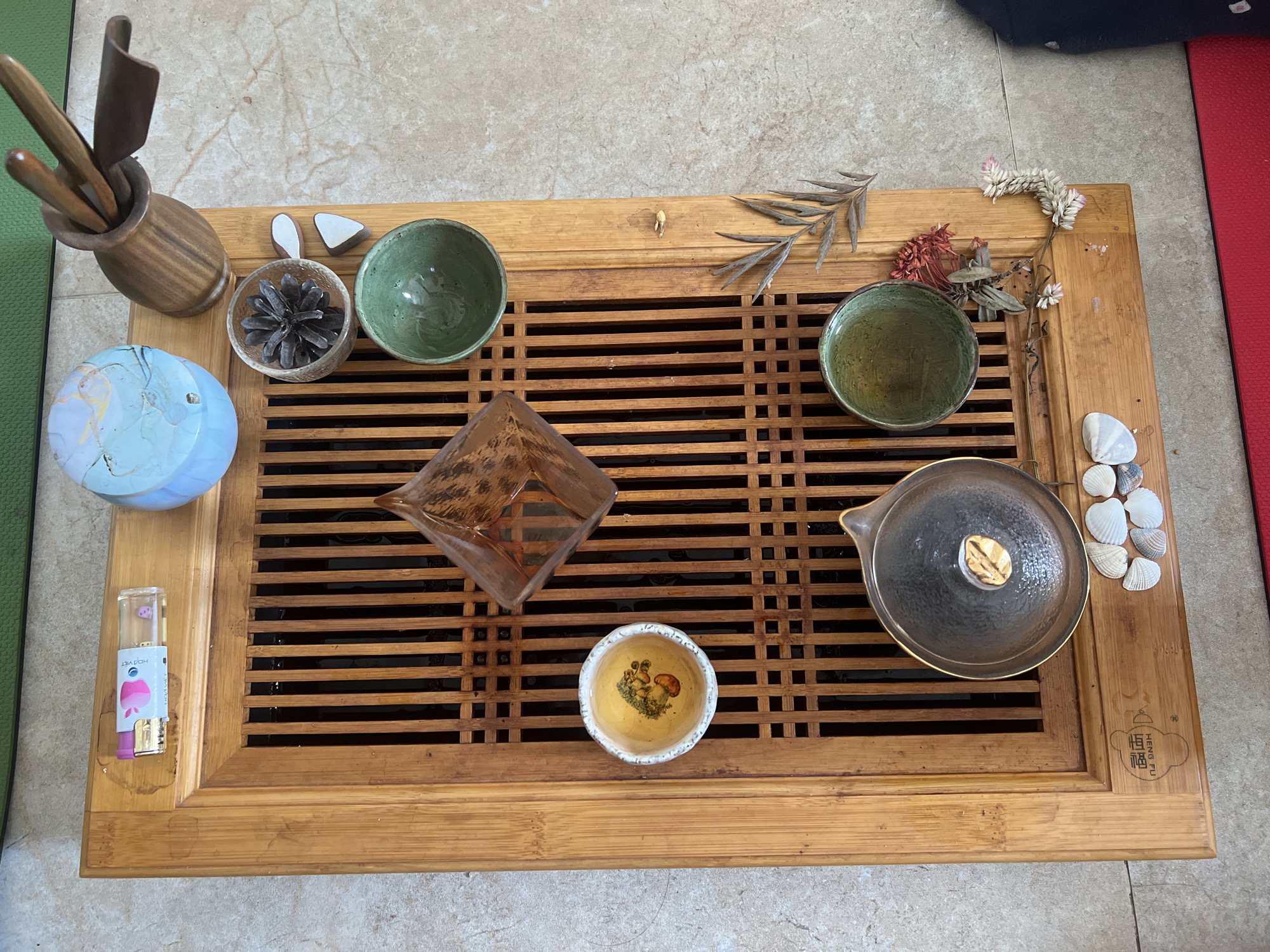A top-down, medium shot captures a bamboo tea tray set on a tiled floor. The tray contains various tea-related items and decorative elements. On the left side of the tray, a small, clear lighter with pink and white accents is visible. Adjacent to it, a small, white ceramic bowl holds a golden liquid with some greenery and what appears to be an artistic depiction of a mushroom or fruit. To the right of this bowl, a geometric, amber-colored glass object is positioned on the slatted part of the tray.
Further to the right, a small, dark wooden holder contains several wooden tea utensils. Above this holder, a pale blue, globe-like object with swirling white and yellow patterns sits on the tray. Below the globe, a small, clear glass bowl contains a dark pinecone. Next to the pinecone are two small, light brown, triangular objects.
Two green ceramic tea bowls are also present on the tray. One is situated below the pinecone, and the other is further down and to the right, closer to the edge of the tray. Near the right edge of the tray, a dried brown leaf rests on the bamboo. Below the tea bowls, a dark, lidded teapot or gaiwan with a gold rim and a gold leaf-shaped knob sits on the slatted surface. To the bottom left of the teapot, a cluster of small white seashells is arranged. The bamboo tray itself has a slatted design and is positioned on a light-colored tiled floor. A hint of a green mat can be seen at the top of the frame, and a red mat at the bottom. The overall scene suggests a tea ceremony setting, possibly in a home or a dedicated space for tea in Da Nang, Vietnam, given the context provided.