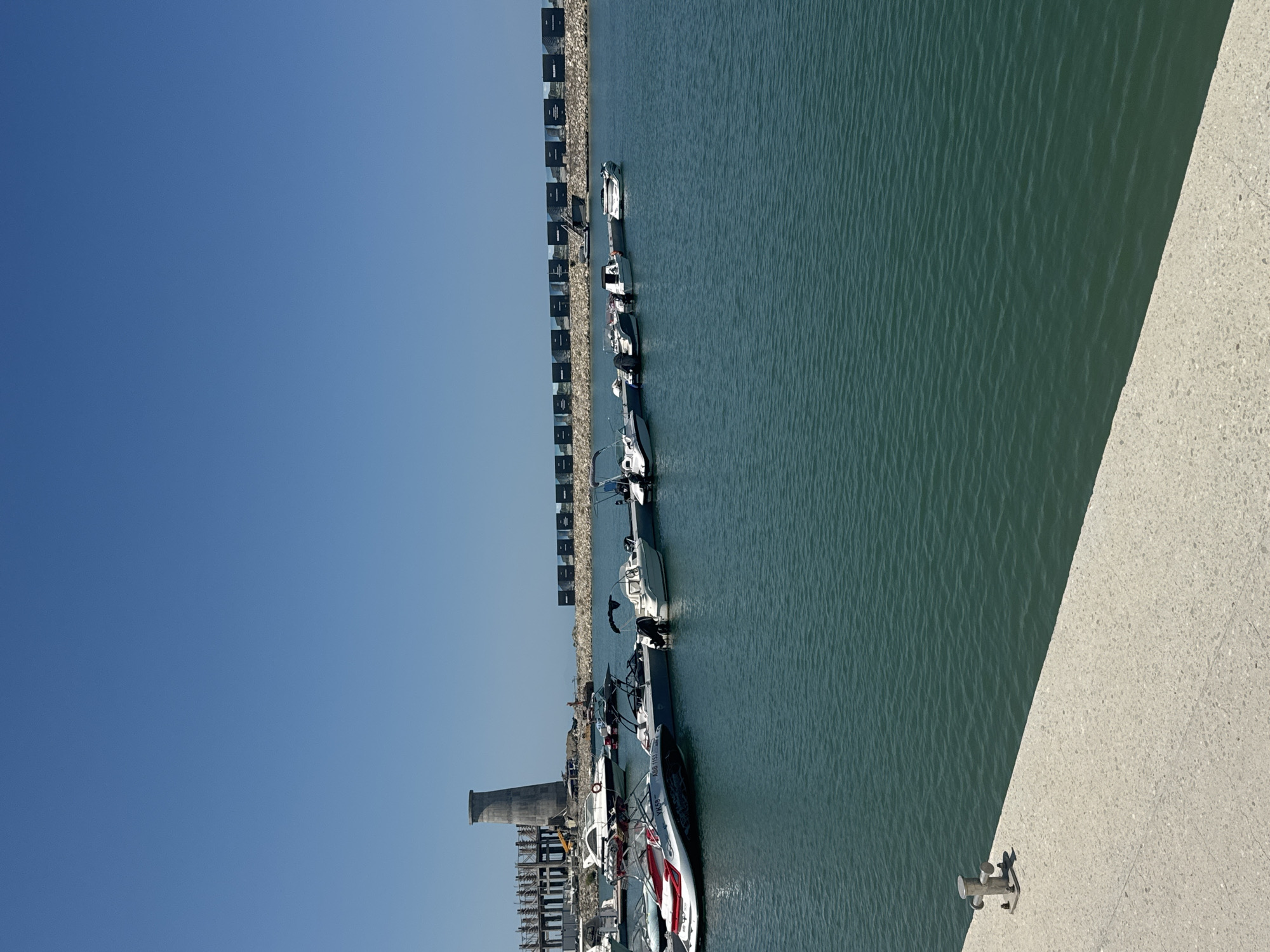The image depicts a view of a marina in Sabunchu Raion, Azerbaijan. The foreground shows a concrete dock with a silver mooring bollard. The water is a deep green color, and several boats are moored in the harbor.
In the middle ground, a line of small motorboats and a jet ski are docked. Further back, a larger white yacht is visible. The background features a stone breakwater topped with a series of rectangular structures, likely shipping containers, arranged in a line. Behind the breakwater and containers, a concrete tower, resembling a lighthouse or industrial chimney, stands prominently against a clear blue sky. The overall scene suggests a sunny day at a coastal location.
