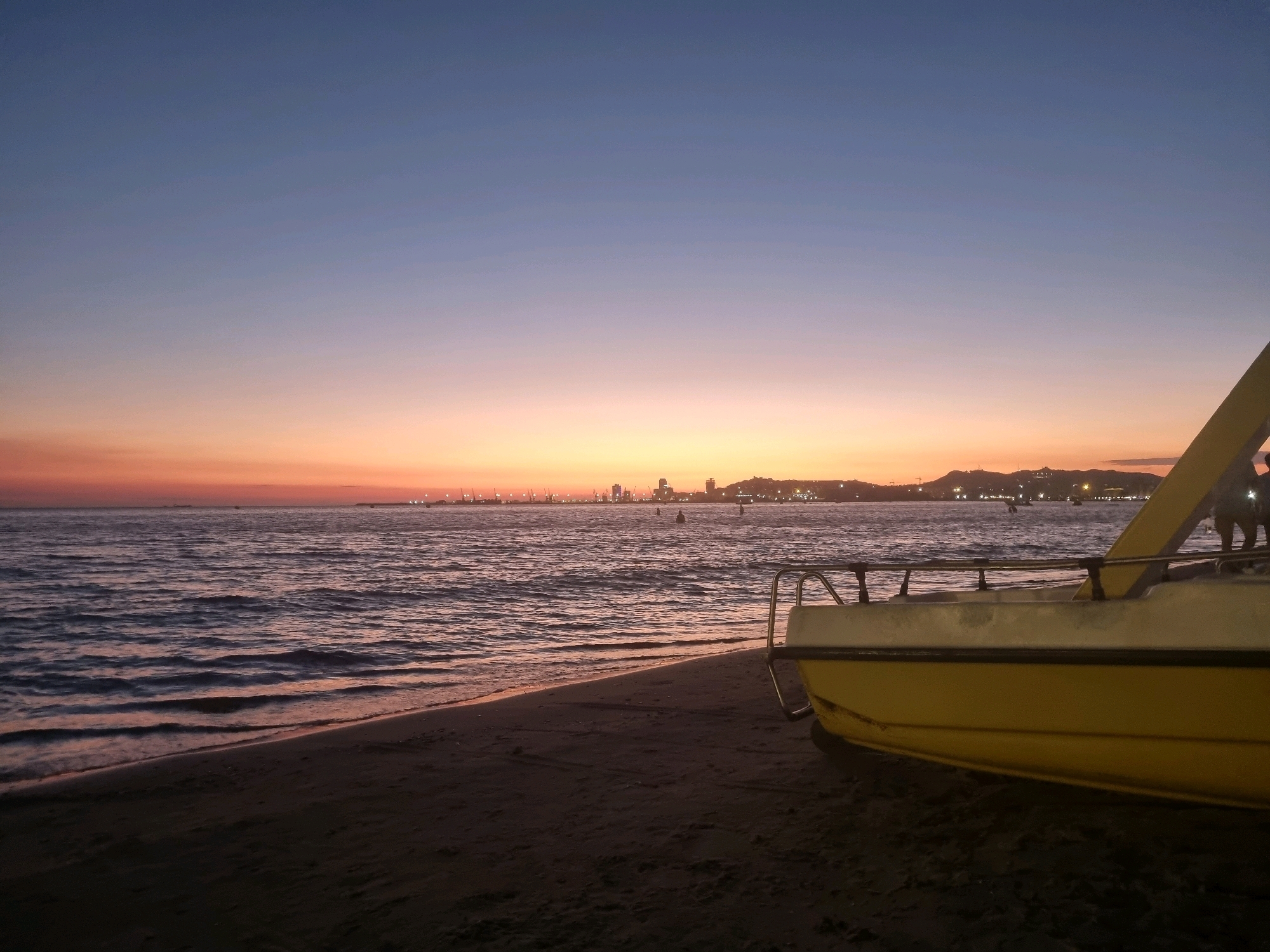 The image depicts a coastal scene at dusk. In the foreground, the bow of a yellow boat rests on a sandy beach. The boat has a metal railing. The sea is calm with gentle waves lapping the shore. In the background, a city skyline is visible on the horizon, with buildings and industrial cranes illuminated by lights. The sky transitions from a deep blue at the top to a soft orange and pink near the horizon, indicative of sunset. The overall scene suggests a quiet evening by the sea. The location is likely the coastline of Bashkia Durrës, Albania, known for its beaches and port city.