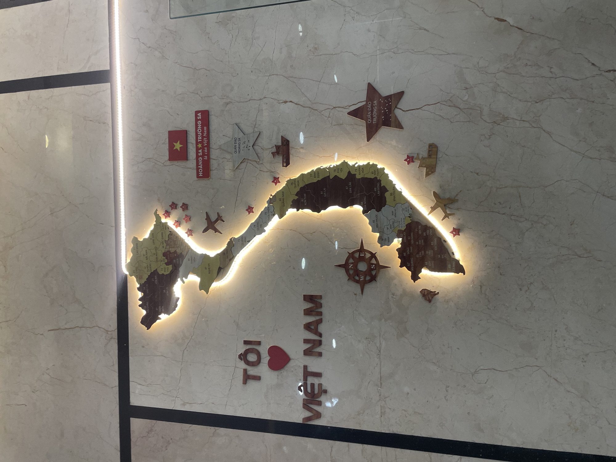 The image shows a decorative, backlit wall map of Vietnam, presented against a light-colored marble wall. The map, illuminated with a warm glow, outlines the distinct S-shape of the country and features internal divisions with various province names and numbers, such as QUẢNG NAM 92, KON TUM 82, GIA LAI, ĐAK LAK 47, and NGHỆ AN 37.
Several wooden cutouts and plaques accompany the map. To the top left, a small Vietnamese flag with a yellow star is placed next to a red rectangular plaque that reads "HOÀNG SA TRƯỜNG SA là của Việt Nam" (Hoang Sa, Truong Sa belong to Vietnam). Two star-shaped wooden plaques are also present: one states "QUÂN ĐẢO HOÀNG SA" (Hoang Sa Archipelago) and the other, "QUẦN ĐẢO TRƯỜNG SA" (Truong Sa Archipelago). Additional decorative elements include several small red star cutouts, two wooden airplane figures, and a wooden compass rose to the right of the map. Below the map, large wooden letters form the phrase "TÔI ❤️ VIỆT NAM" (I Love Vietnam). A bright vertical light strip runs along the left side of the display. The overall installation conveys a strong message of national identity and territorial sovereignty.