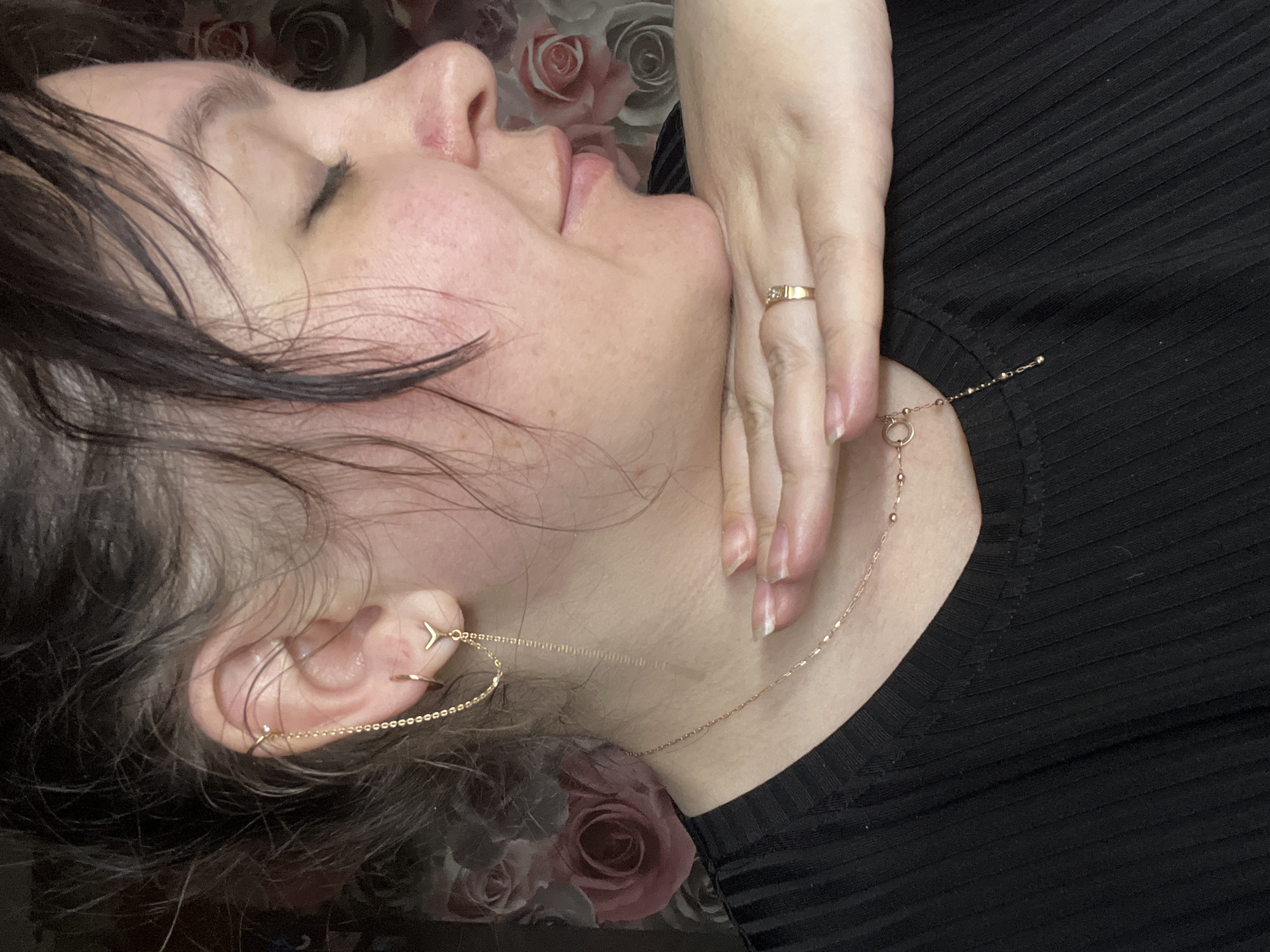 A profile view of an individual, likely female, is captured from the chest up within an indoor setting in Noviy, Russia. The individual's eyes are closed, and their right hand is positioned beneath their chin, providing support. Dark, seemingly damp hair is styled loosely around the face, obscuring part of the forehead and left ear. The skin is fair, exhibiting minor redness on the cheeks and a small, red mark on the nasal bridge. The individual wears a black, ribbed garment. On the visible left ear, a series of gold-toned piercings are present: a small clear stud in the upper helix, a small hoop in the earlobe, and a V-shaped stud below the hoop. A delicate chain connects the helix stud to the V-shaped stud, with an additional longer chain dangling from the V-shaped stud. A thin, gold-toned necklace with small bead-like embellishments and a circular charm with a descending beaded chain is visible around the neck. A gold-toned ring, featuring a small square-cut clear stone, is worn on the ring finger of the supporting right hand. The background consists of a dark wall covering with a repeating pattern of large, stylized pink and red roses.